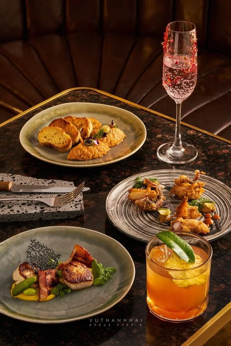 A gourmet meal with a glass of sparkling beverage, featuring a plate of fried seafood, a plate of seared meat with vegetables, a plate of bread with spreads, and a glass of orange-colored cocktail garnished with lemon and mint leaves, on a dark marble table.