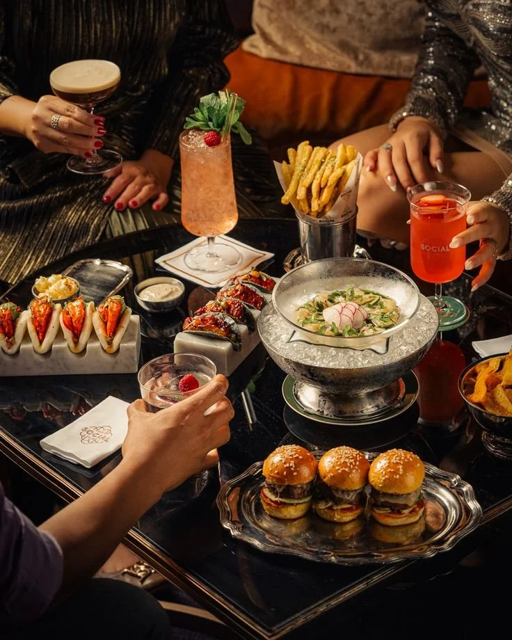 A table with various dishes and drinks, including sliders, sushi, a bowl of pasta, French fries, and colorful cocktails, with people reaching for food and drinks at a social gathering.