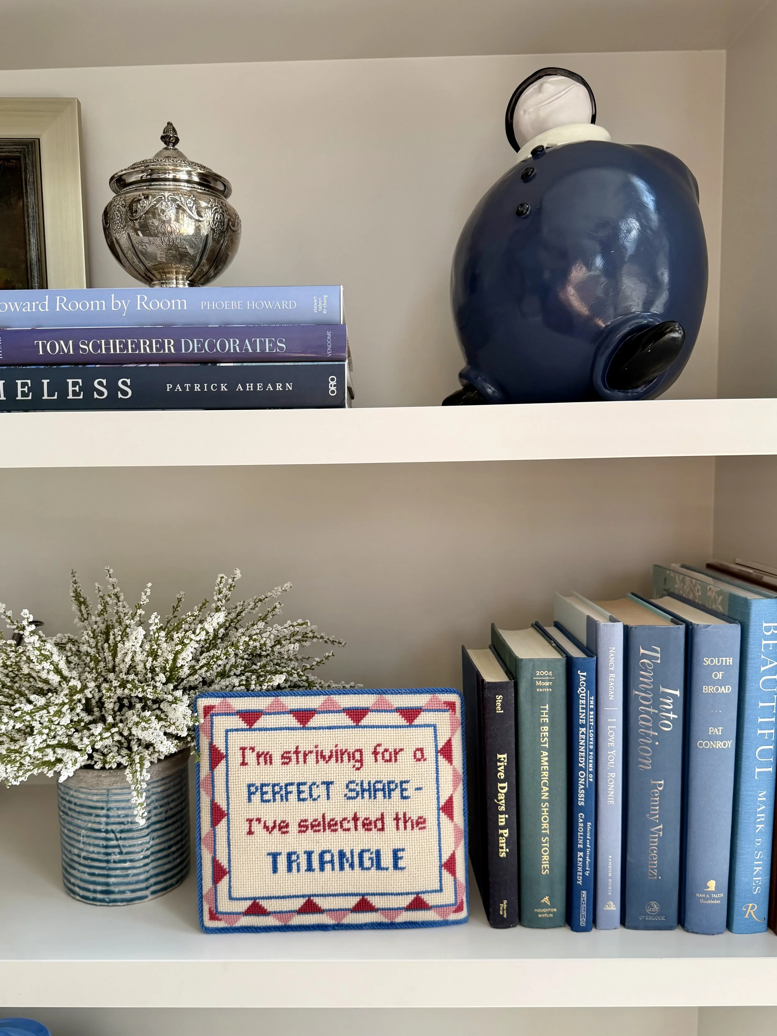 A white bookshelf holding a blue and white vase with white flowers, a stack of books including titles like 'How to Talk to Americans,' 'Five Days in Paris,' and 'The Best American Short Stories,' and a decorative embroidered sign that reads 'I’m striving for a perfect shape - I’ve selected the triangle.' The top shelf also has a silver decorative jar and a large blue teapot-shaped figurine.