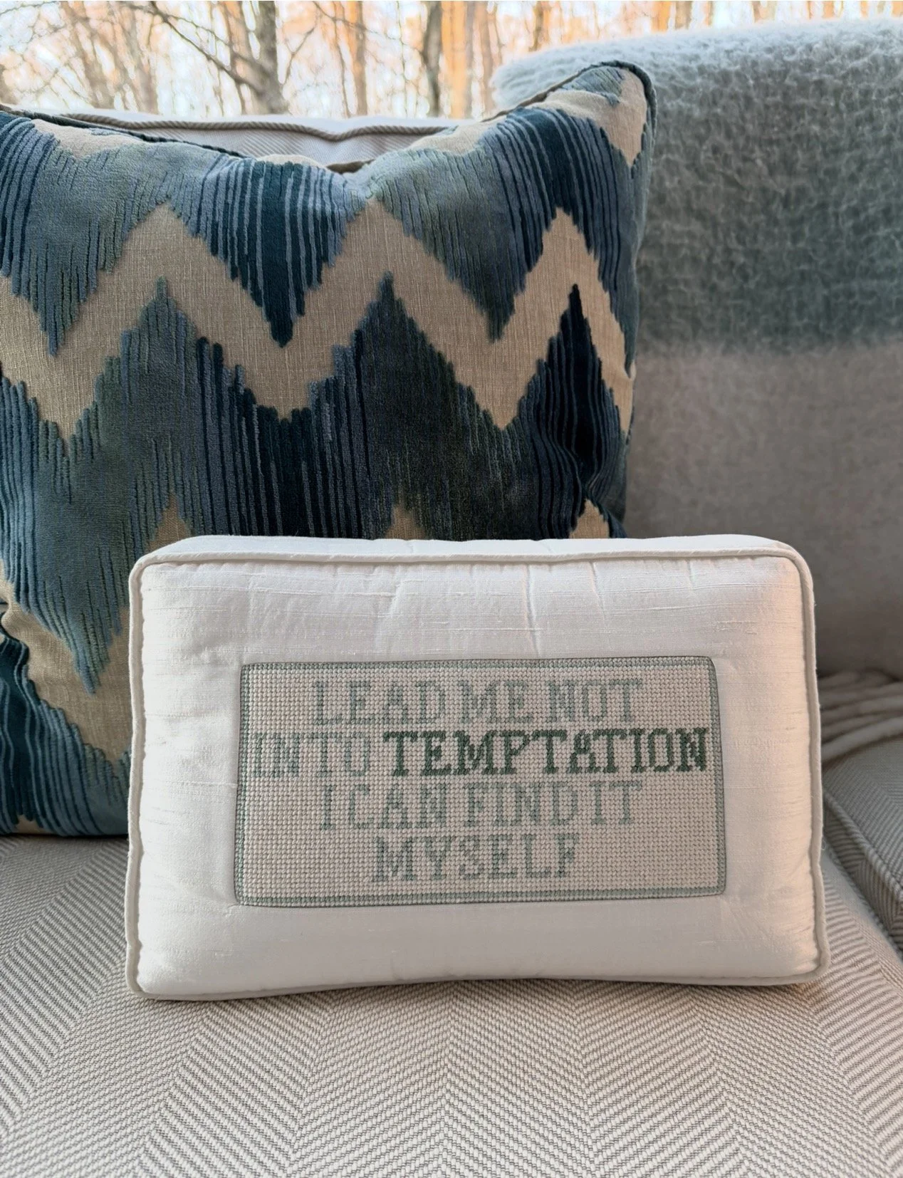 A decorative pillow with a woven message that reads, 'LEAD ME NOT INTO TEMPTATION, I CAN FIND IT MYSELF,' standing in front of a larger patterned pillow on a couch.