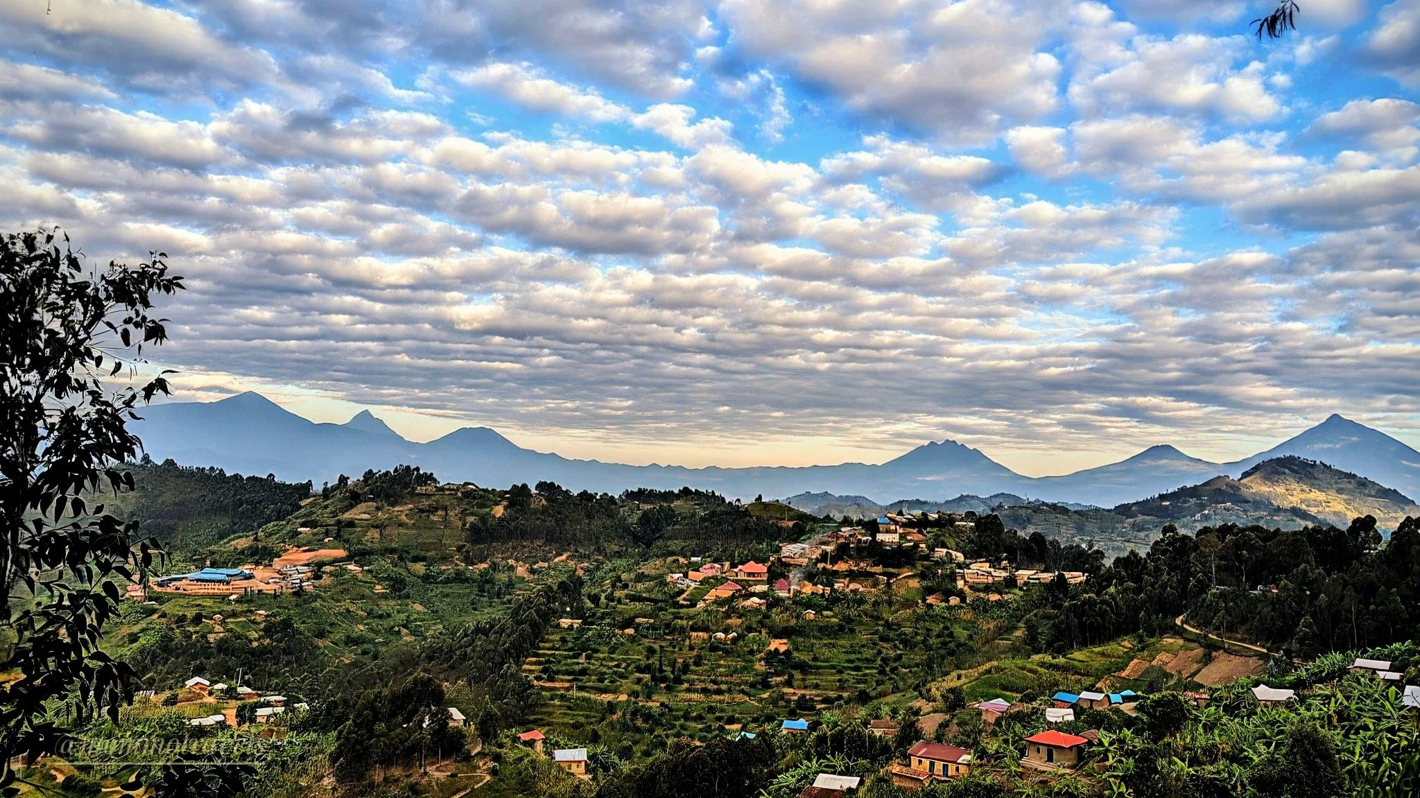Scenic view of a mountainous landscape with a small village, lush green hills, and a sky filled with scattered clouds.