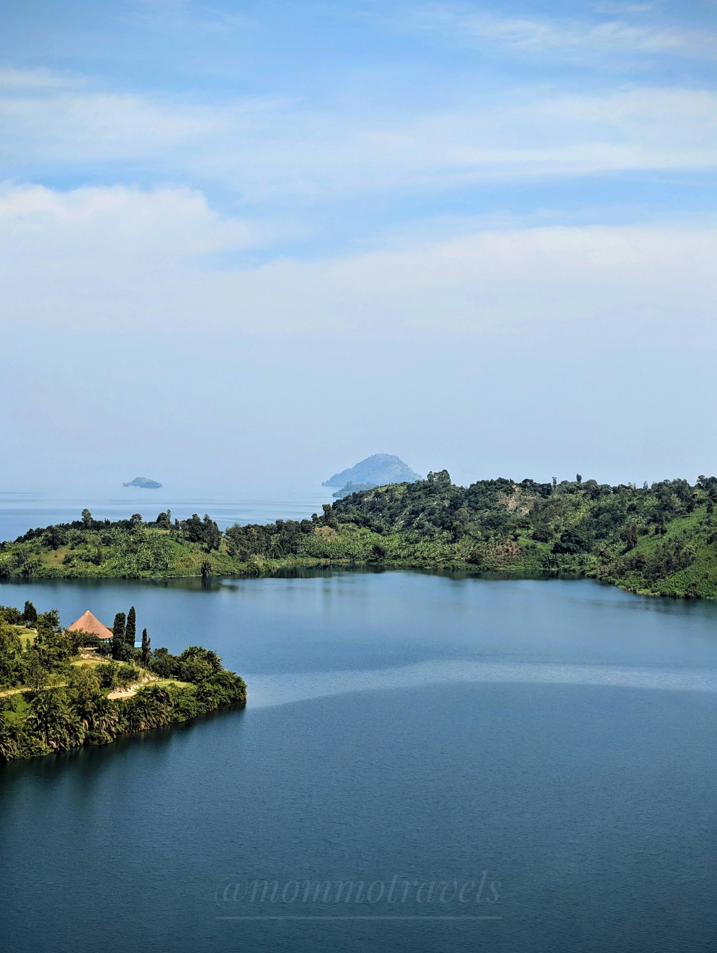 A scenic view of a large lake with a lush green shoreline and trees, surrounded by rolling hills and mountains under a cloudy sky.