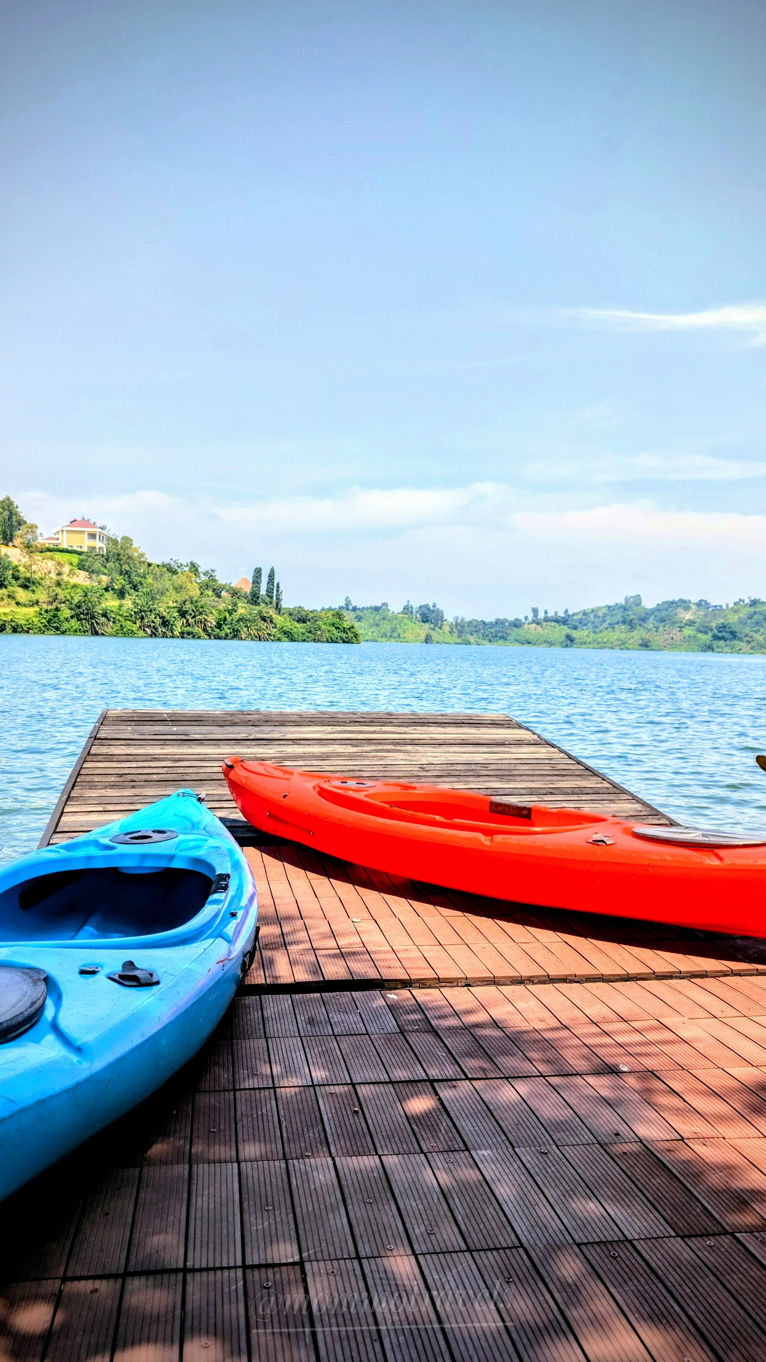 Two kayaks, one blue and one red, resting on a wooden dock by a lake with green hills in the background and a clear blue sky overhead.