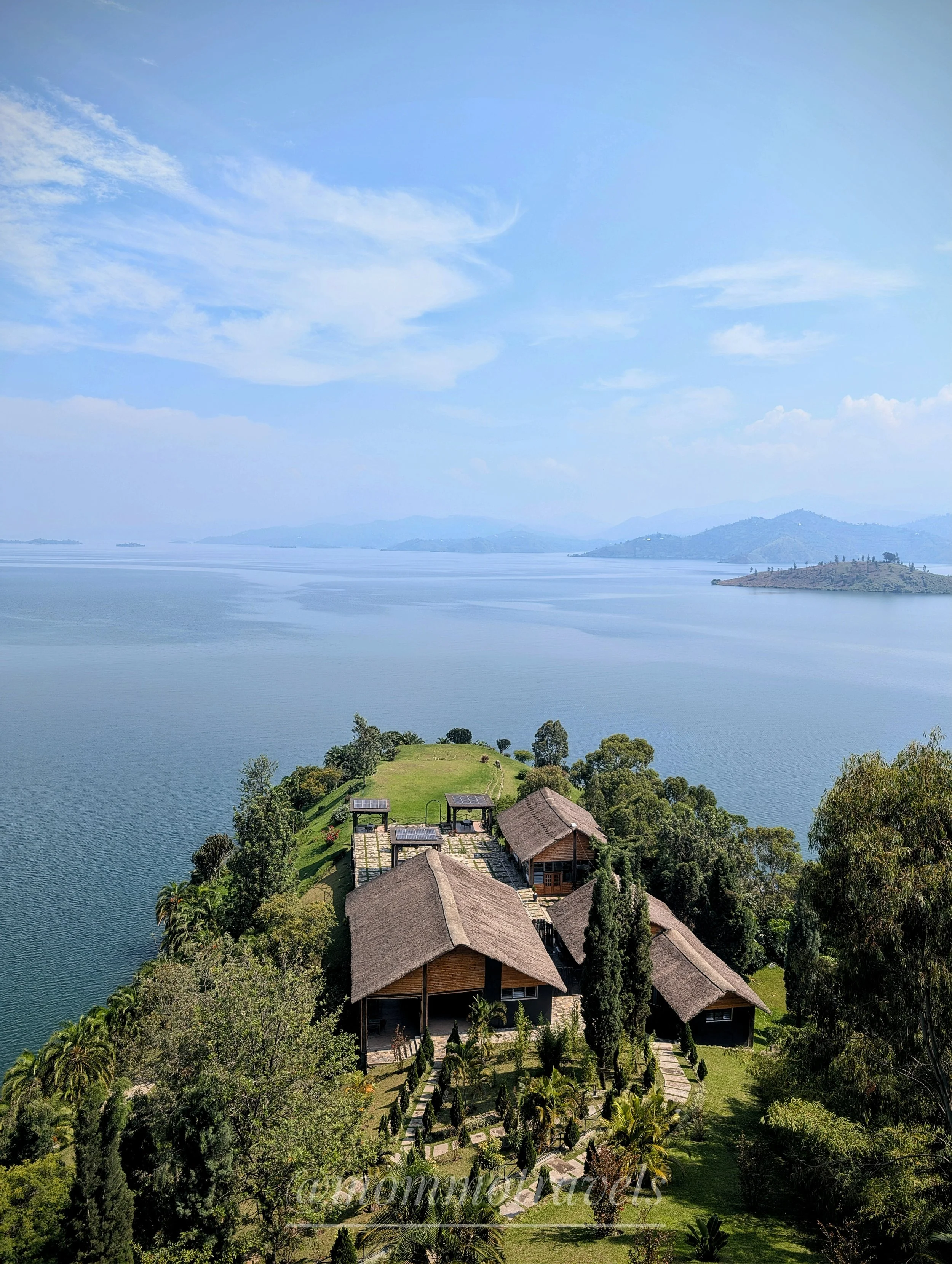 Aerial view of a lakeside with traditional-style houses, lush green trees, and a pier, surrounded by a large body of water and distant mountains under a partly cloudy sky.