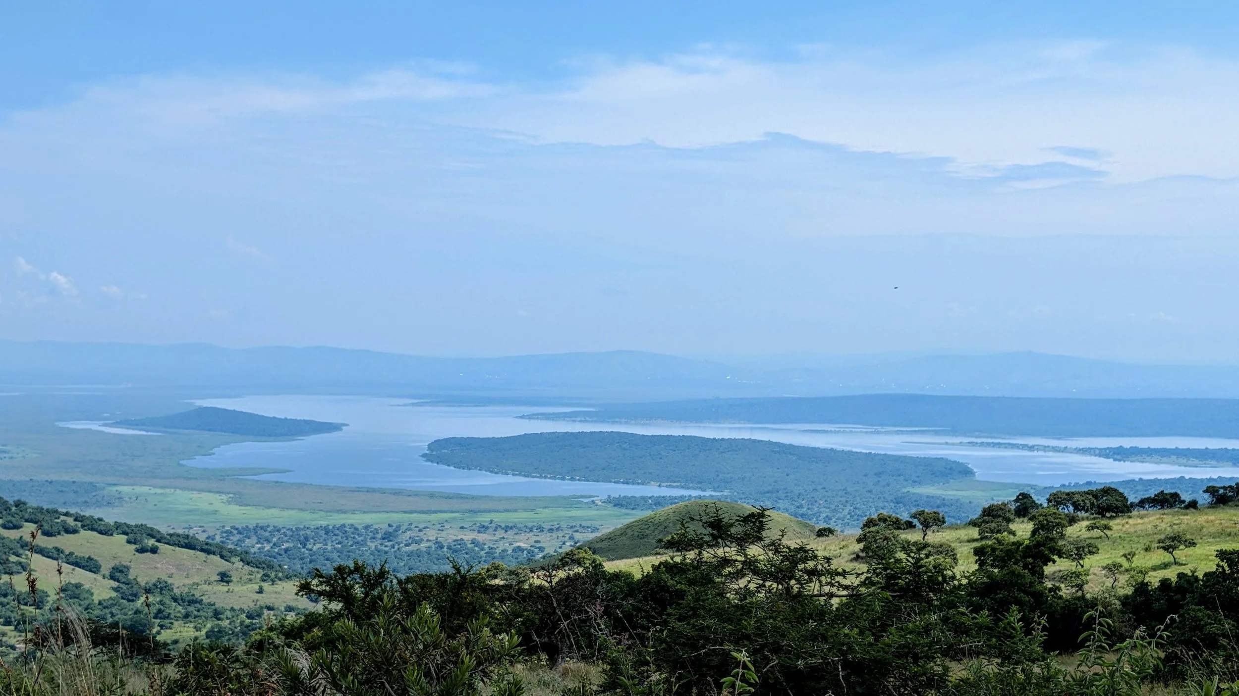 Landscape view of a river winding through green fields and hills, with a backdrop of distant mountains and a partly cloudy sky.