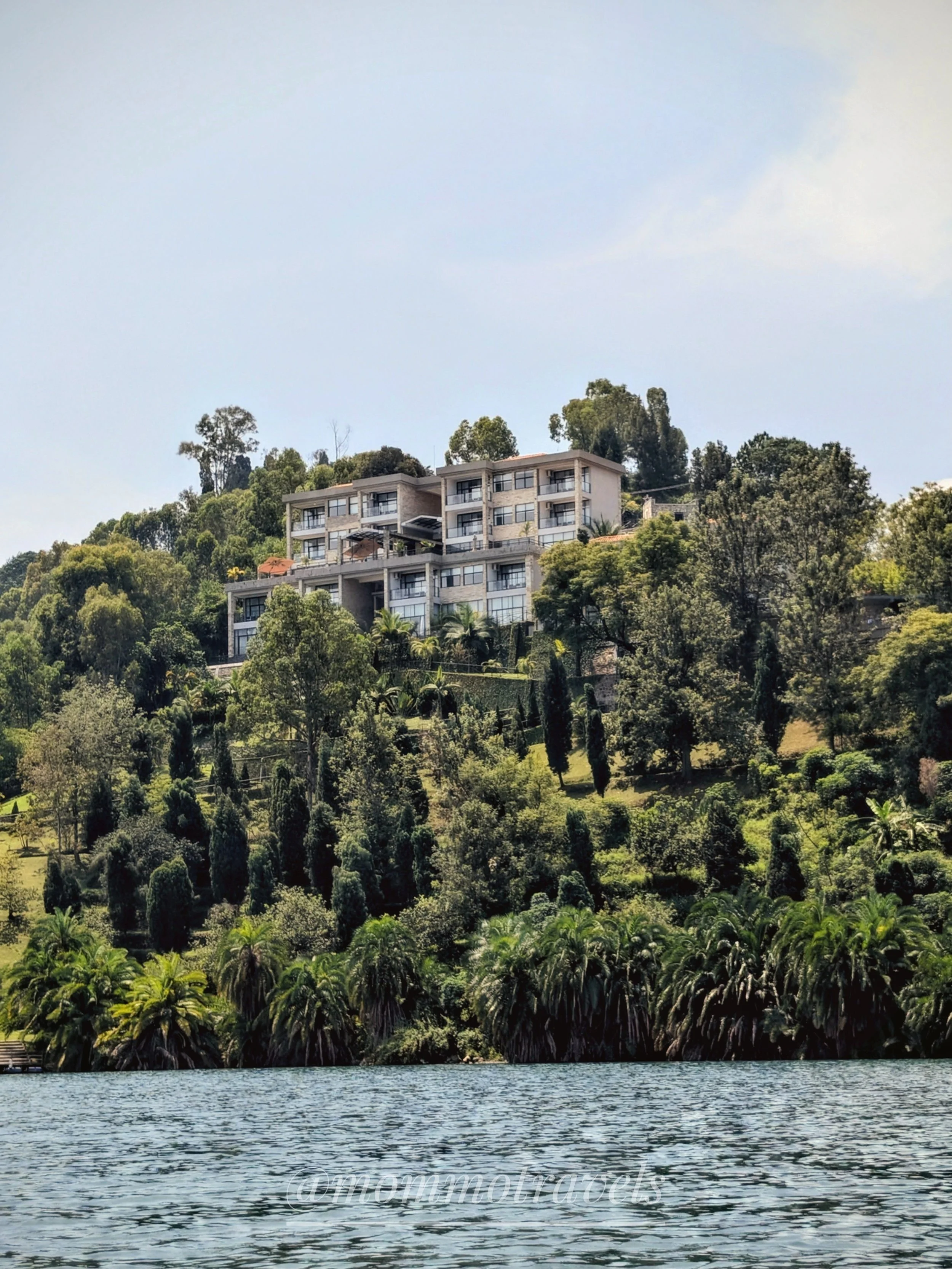Residential buildings on a hillside surrounded by lush green trees and vegetation, with a body of water in the foreground and a clear sky above.