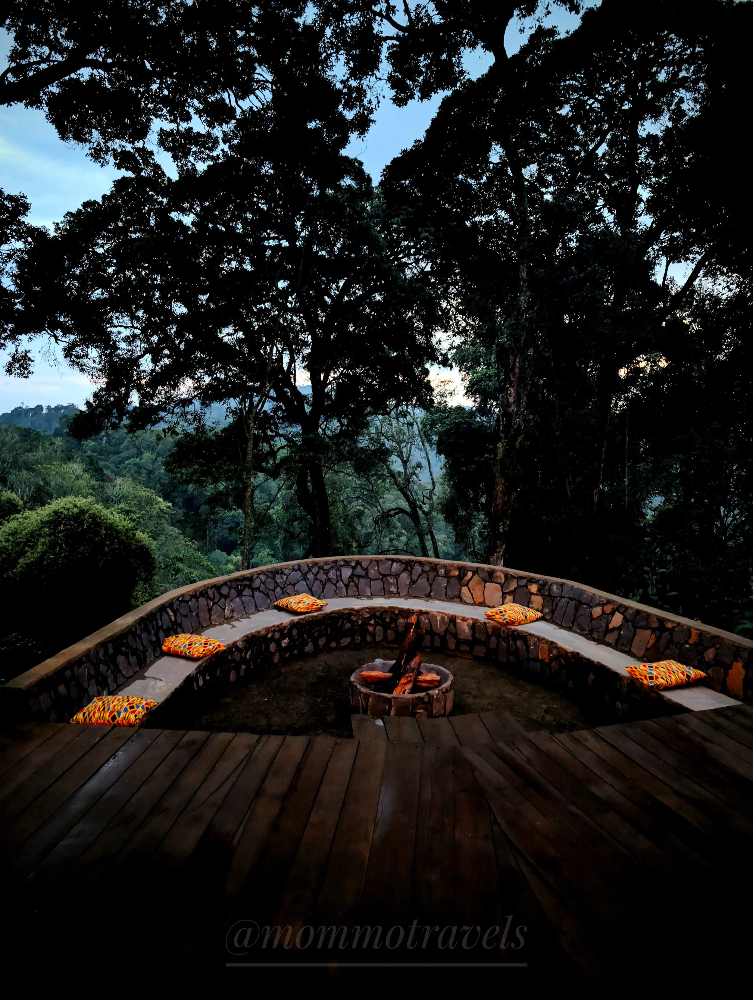 A circular stone seating area with colorful cushions surrounds a fire pit, set outdoors against a backdrop of tall trees and a partly cloudy sky during dusk.