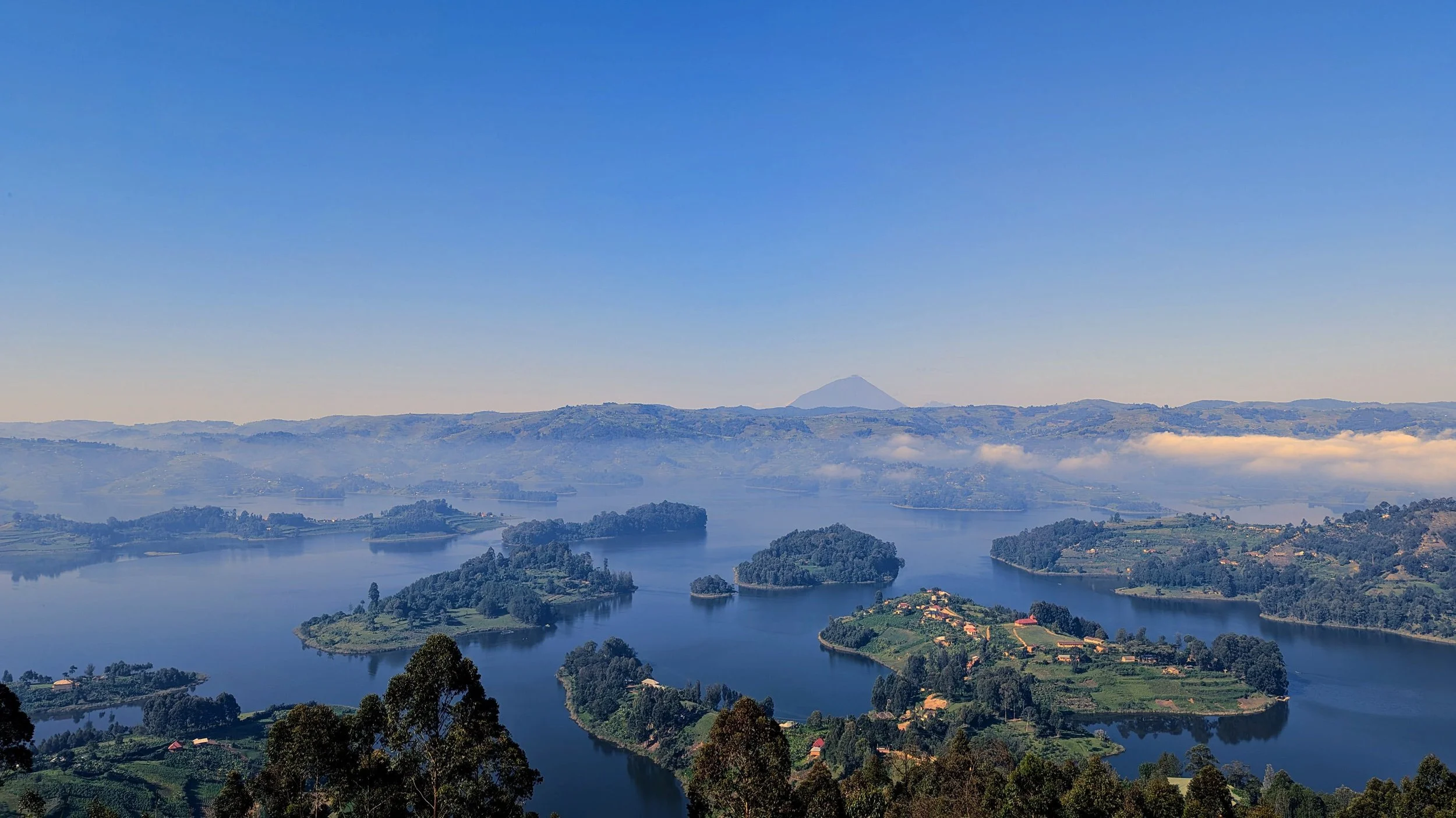 A panoramic view of a large lake surrounded by lush green hills, small islands, and scattered houses, with a distant mountain peak under a clear blue sky.