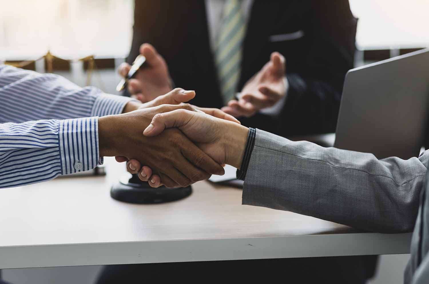 Two individuals are shaking hands across a conference table while others in the background discuss, with a laptop and microphones on the table.