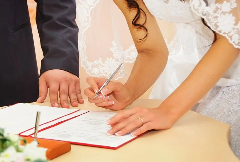 Bride signing a marriage certificate with groom's hand on the table