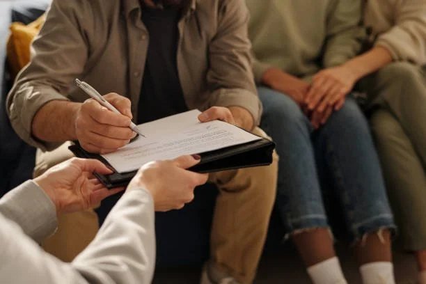 Two people sitting on a couch, with one writing on a clipboard held by a third person.