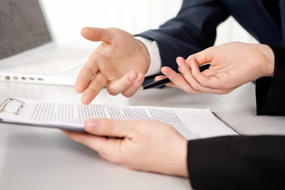 Two people in business attire discussing a document at a desk, one person pointing at the paper.