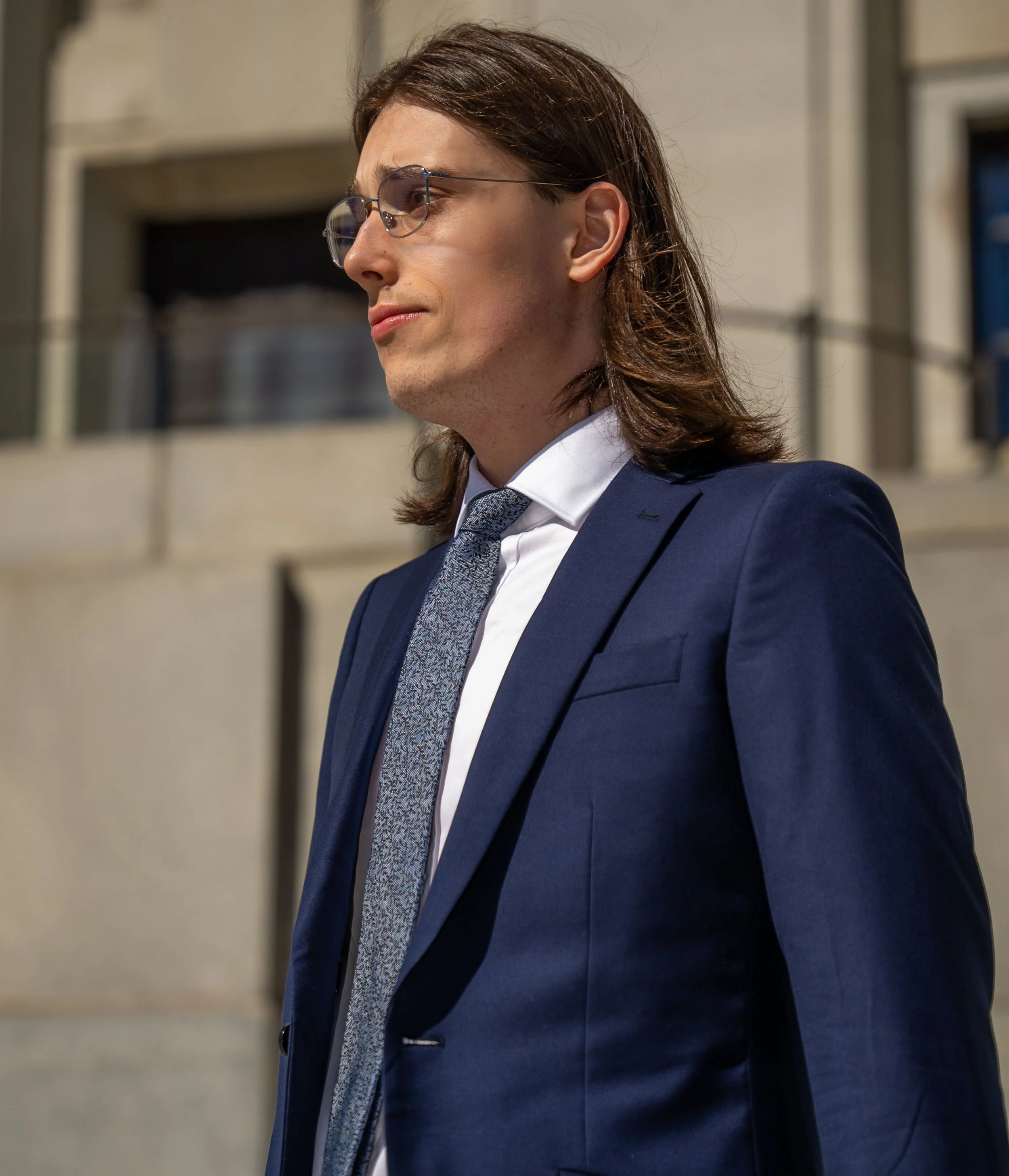 A side profile of a young woman with long brown hair wearing glasses and a dark blue business suit with a patterned tie, standing outdoors in an urban setting.