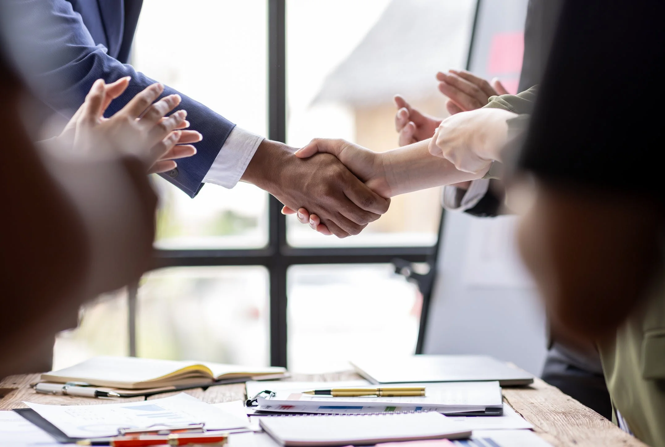 People shaking hands over a table in a professional setting, with notebooks and papers on the table representing a business or meeting environment.