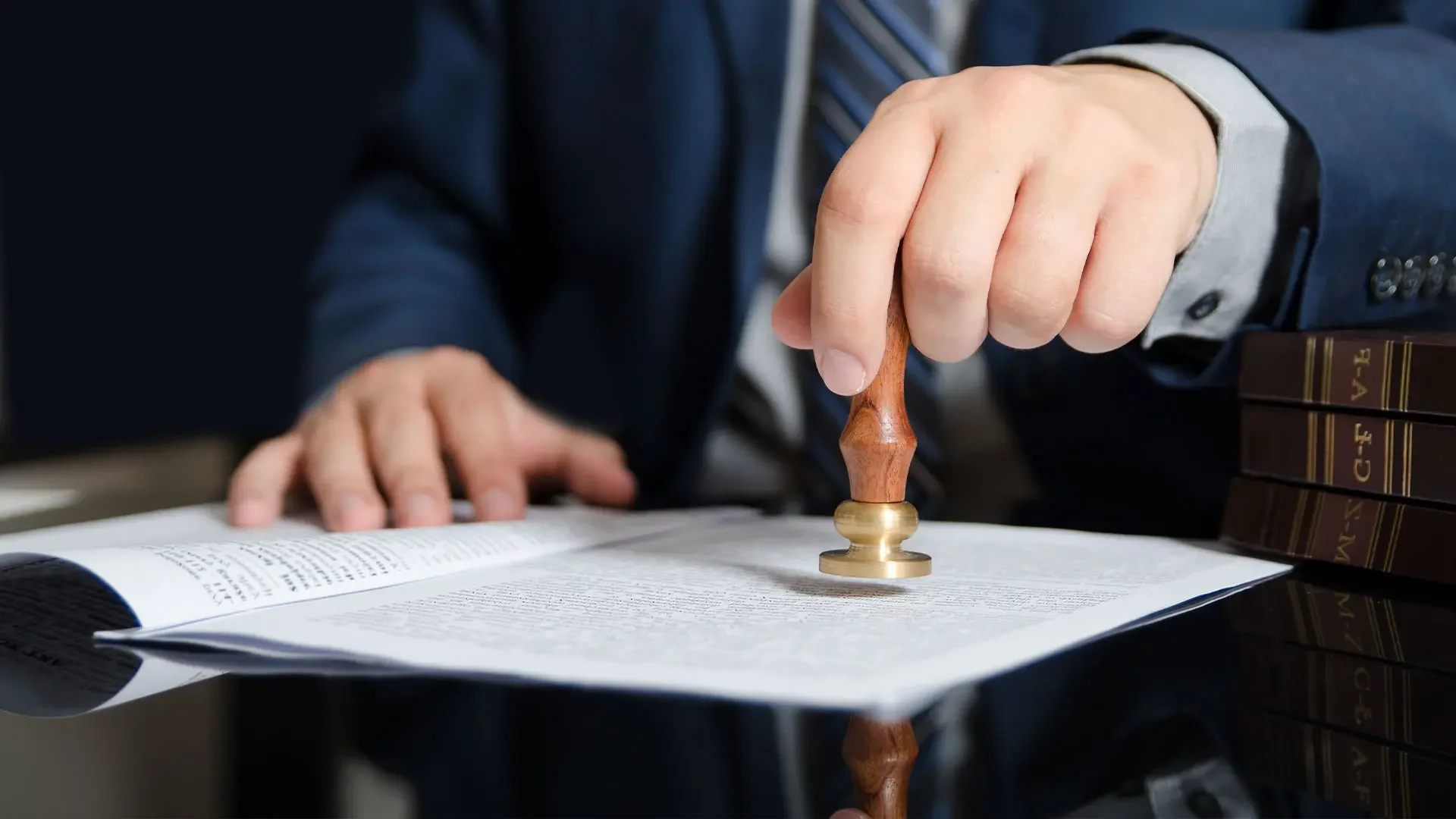 A person in a business suit stamping a document with a wooden seal. There are legal books stacked on the table.