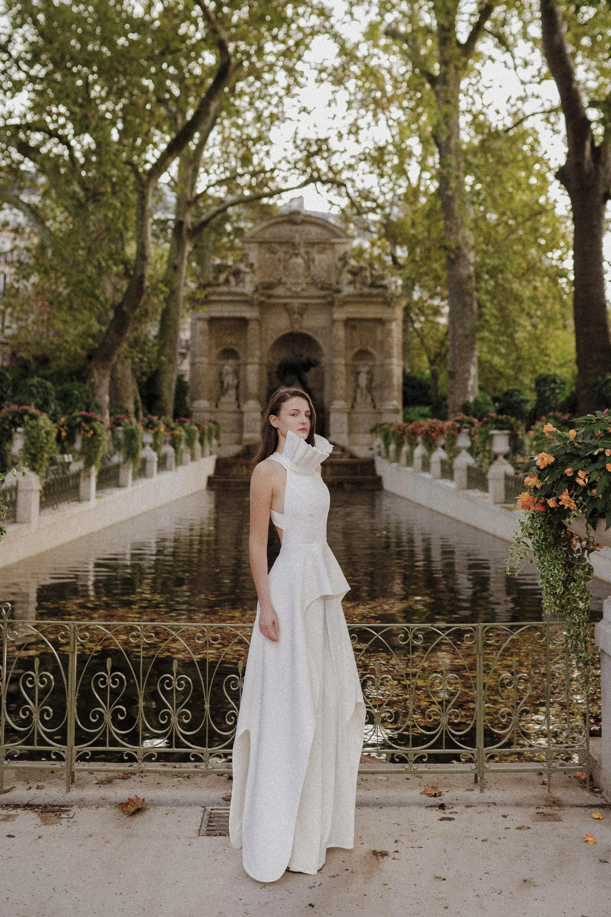 Una mujer con vestido largo y blanco, de cabello castaño, que está parada frente a una cerca de metal en un parque con árboles y flores en la época de otoño.