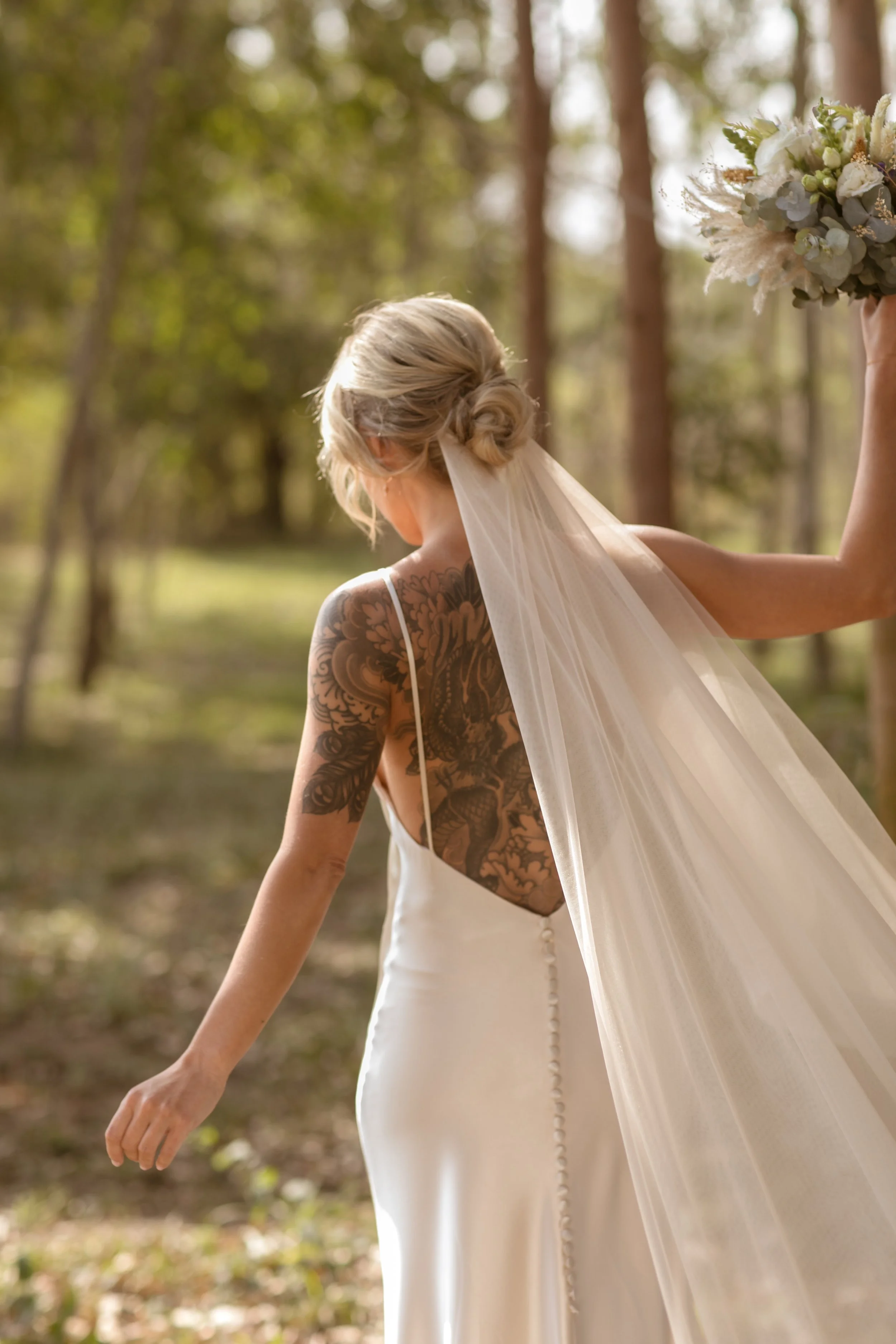 Mujer con cabello rubio recogido, vestido blanco y sombra de tatuajes en el brazo, en un bosque, sosteniendo un ramo de flores blancas y verdes, con un velo largo.