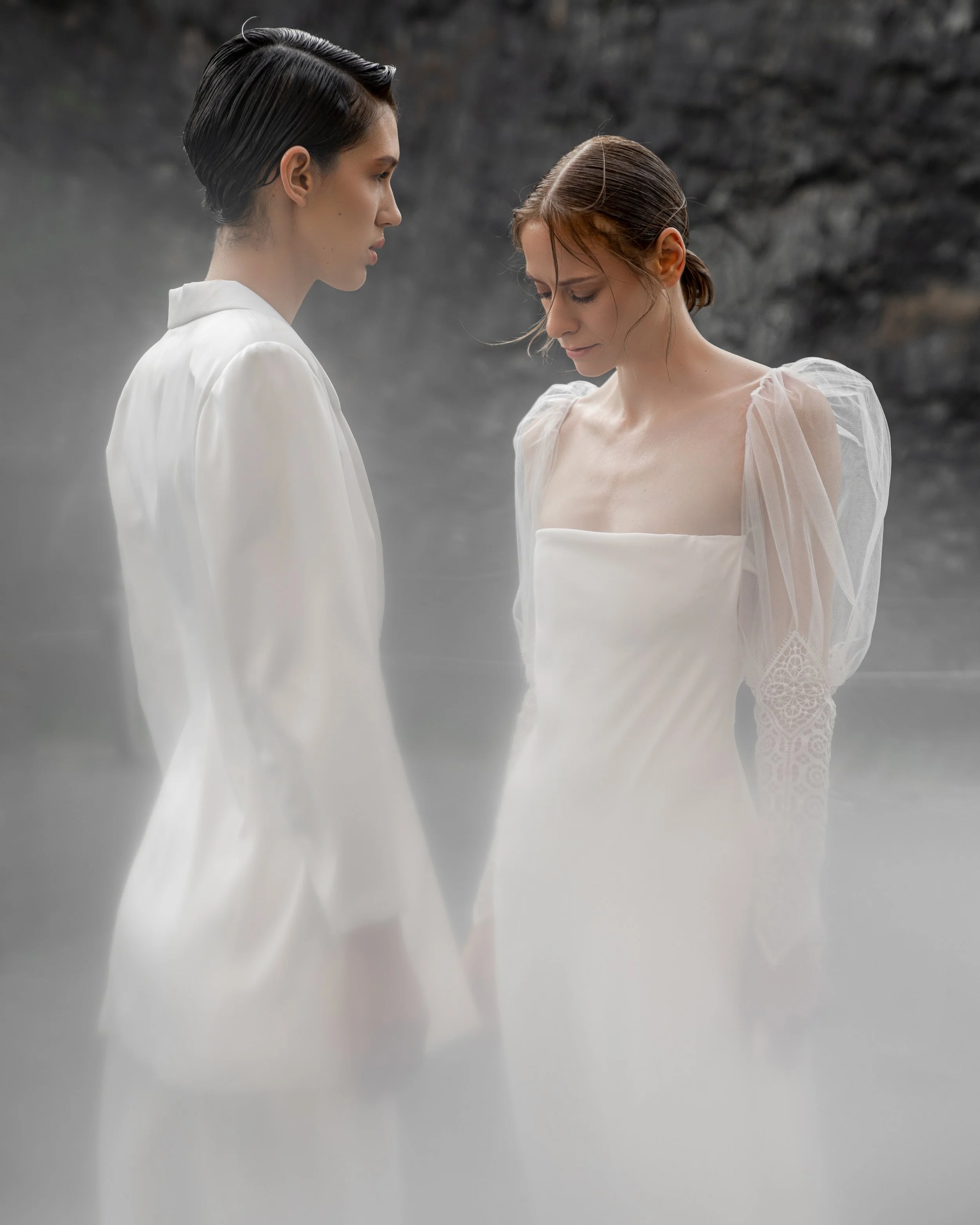 Dos novias, una con traje blanco y la otra con vestido de mangas abullonadas (ambas piezas de Ofelia Otello). En un entorno hostil sobrevive el amor. Fotografía de Nath Planás.