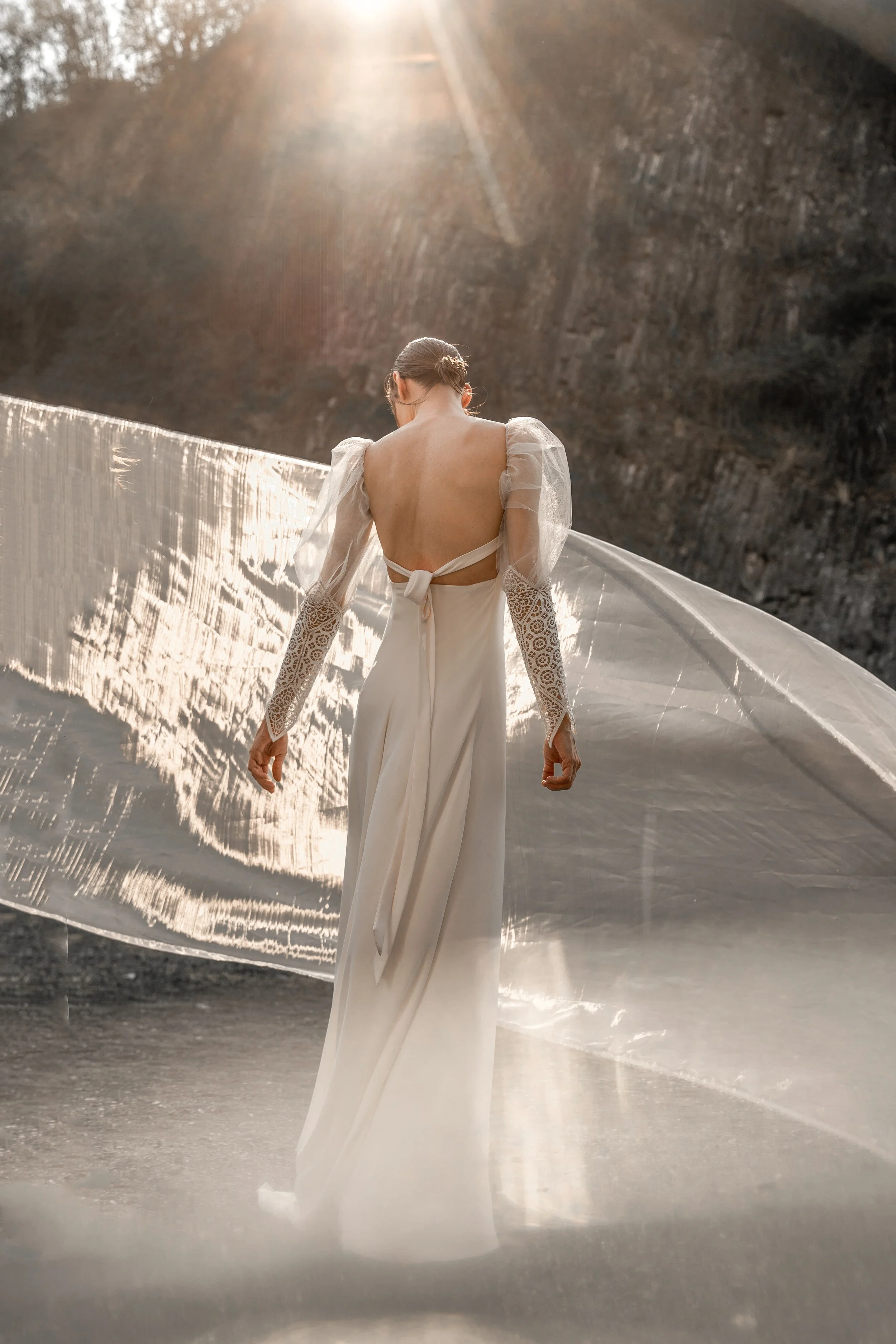 Mujer con vestido blanco, de espaldas, en un entorno natural con sol brillante y montañas. Vestido de Ofelia Otello. Fotografía de Nath Planás.