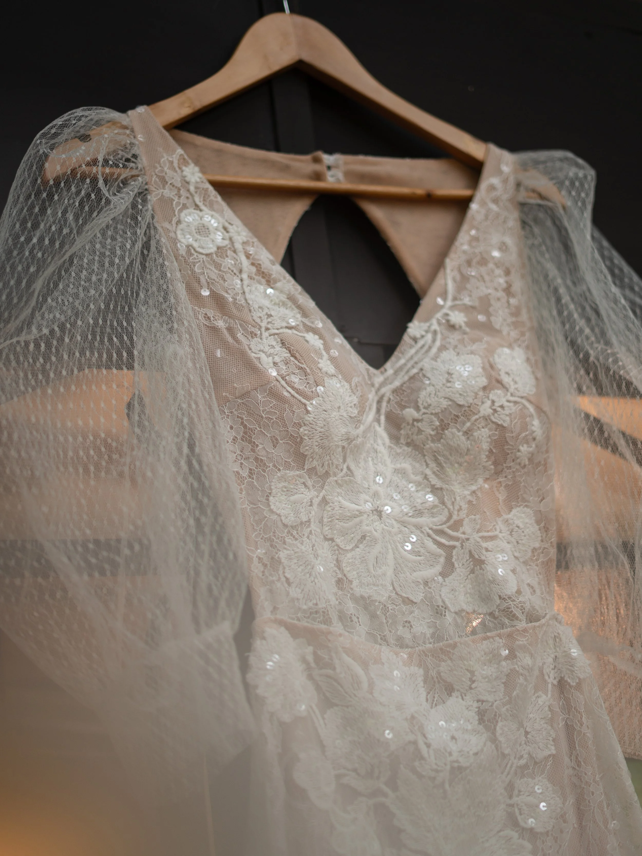 Vestido de novia de encaje con detalles de hilo y lentejuelas, colgado en una percha de madera, en un fondo oscuro.