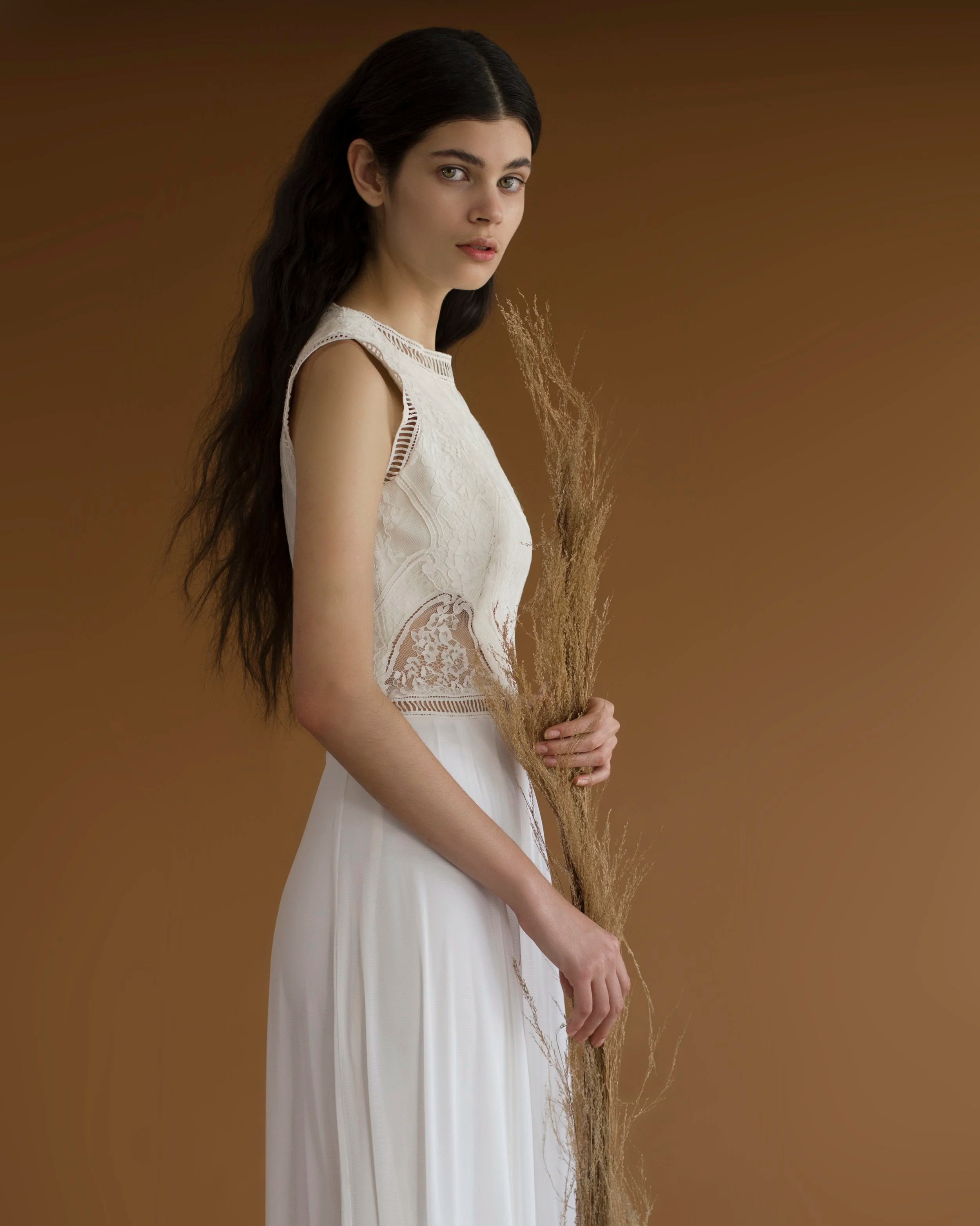 Joven mujer con cabello largo y oscuro, vestida con blusa blanca de encaje y pantalones blancos, sosteniendo un ramo de orozuz, en un fondo marrón.