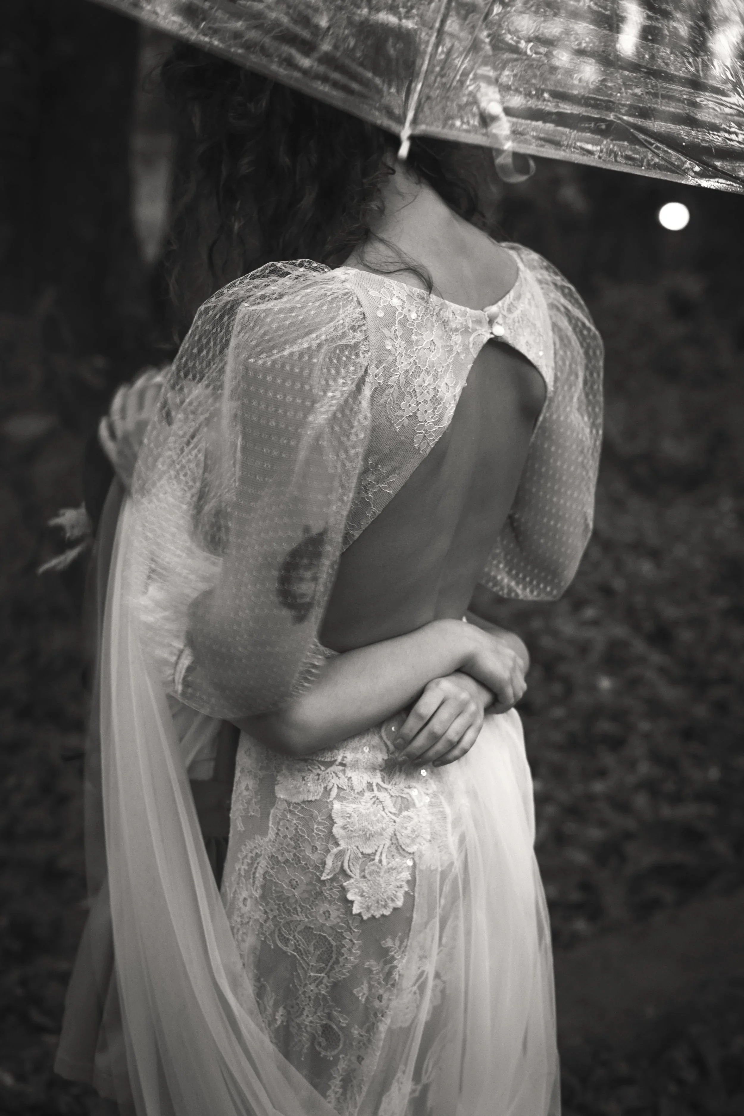 Mujer abrazada a su hija con vestido de encaje y velo de Ofelia Otello, vista de espaldas, con paraguas en un entorno lluvioso. Fotografía de Nath Planás.
