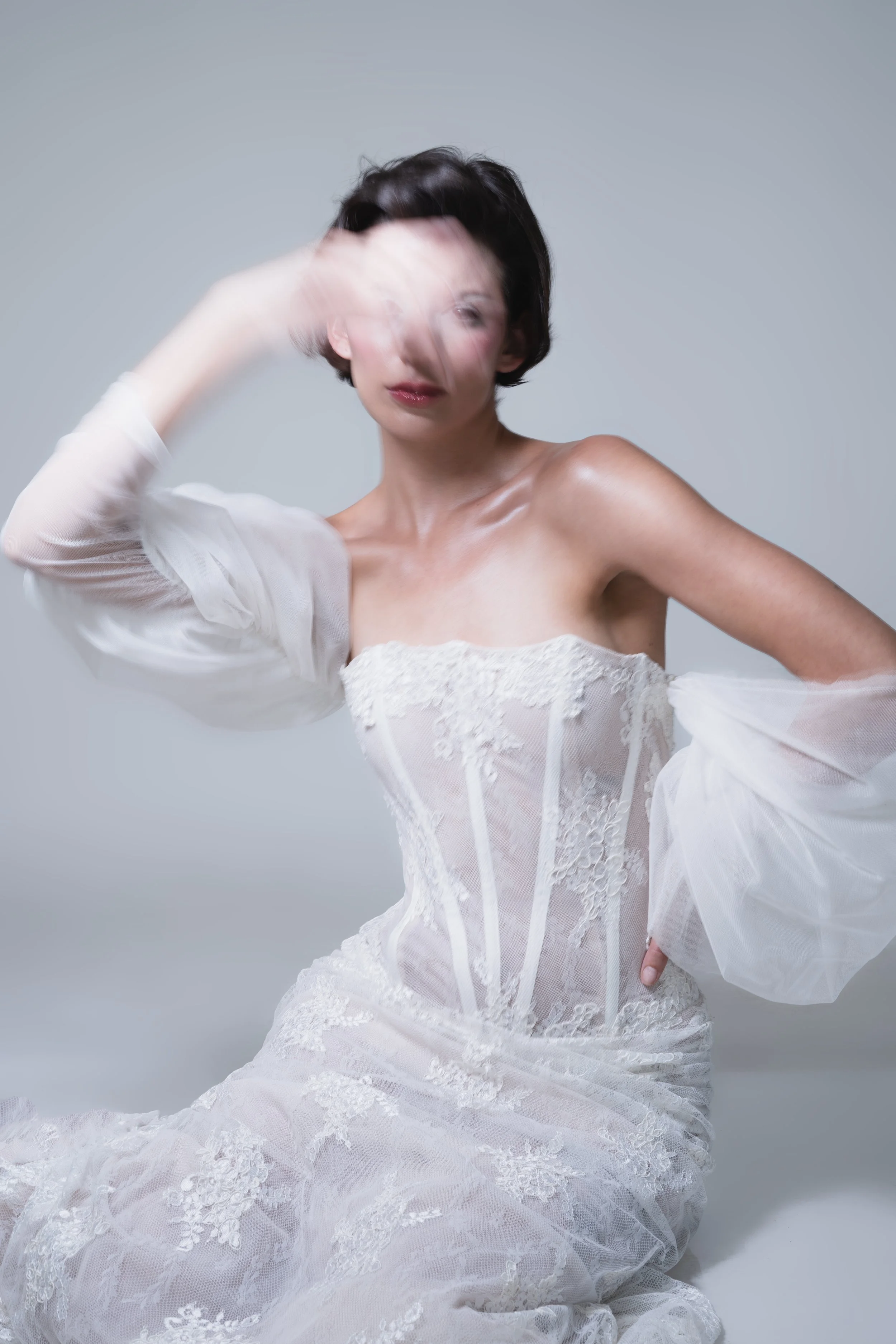 Una mujer con cabello corto, usando un vestido de encaje blanco con mangas de tul de Ofelia Otello, posando con una expresión que su mano no deja ver. Fotografía de Nath Planás.