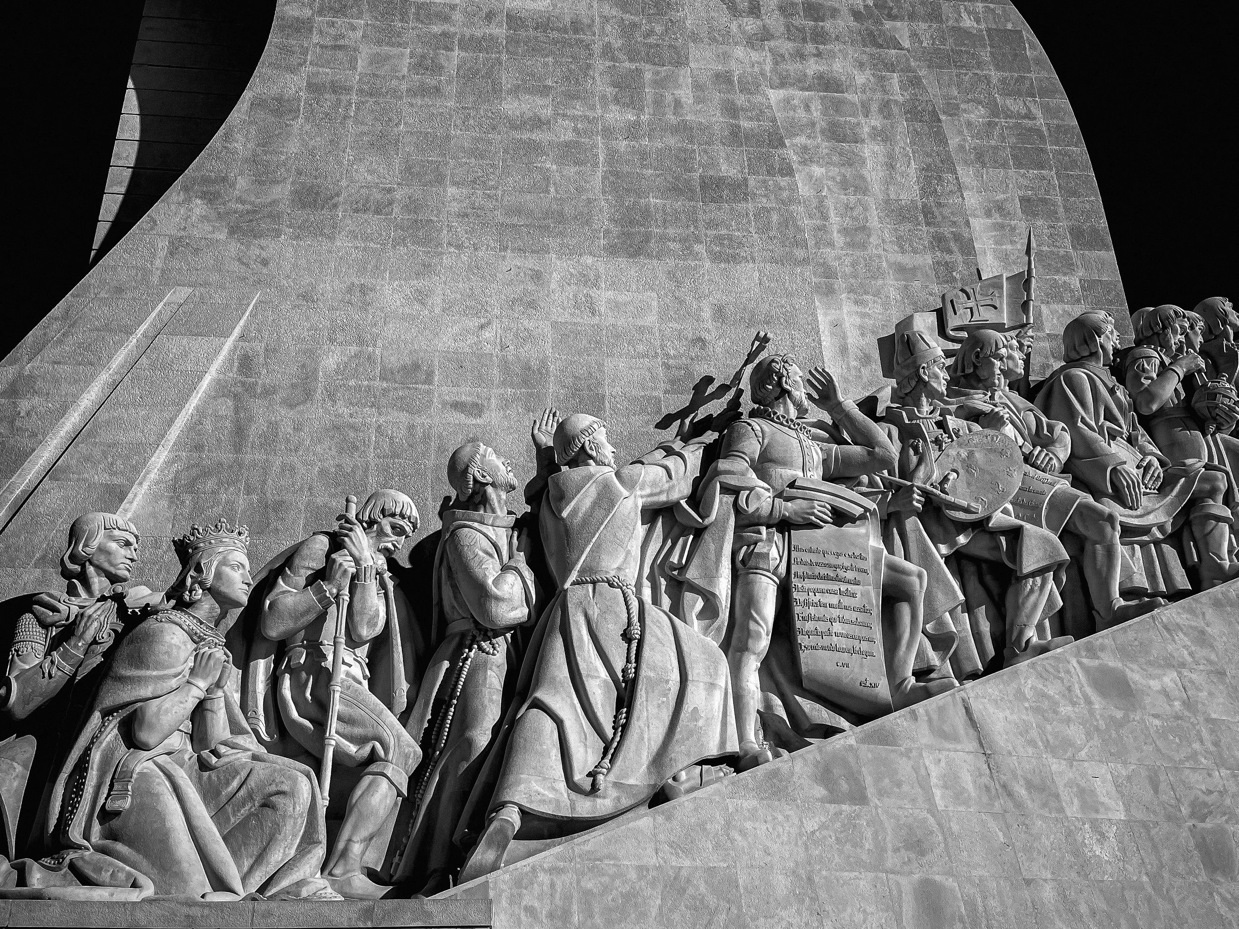 Black-and-white photo of a stone monument featuring a detailed relief sculpture of historical figures in armor, some holding shields and banners.