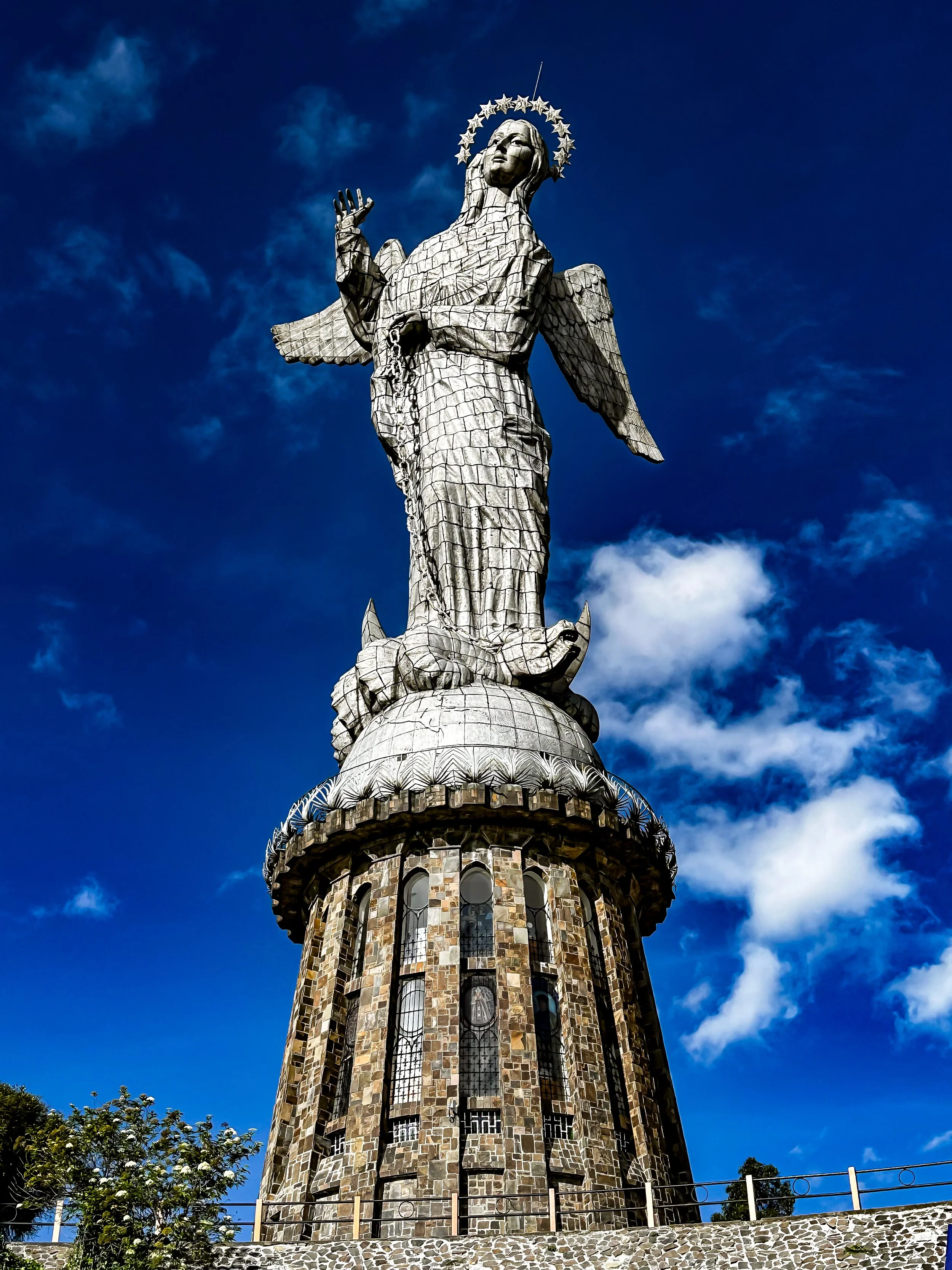 Tall monument featuring a statue of an angel with a halo of stars, set against a blue sky with some clouds.