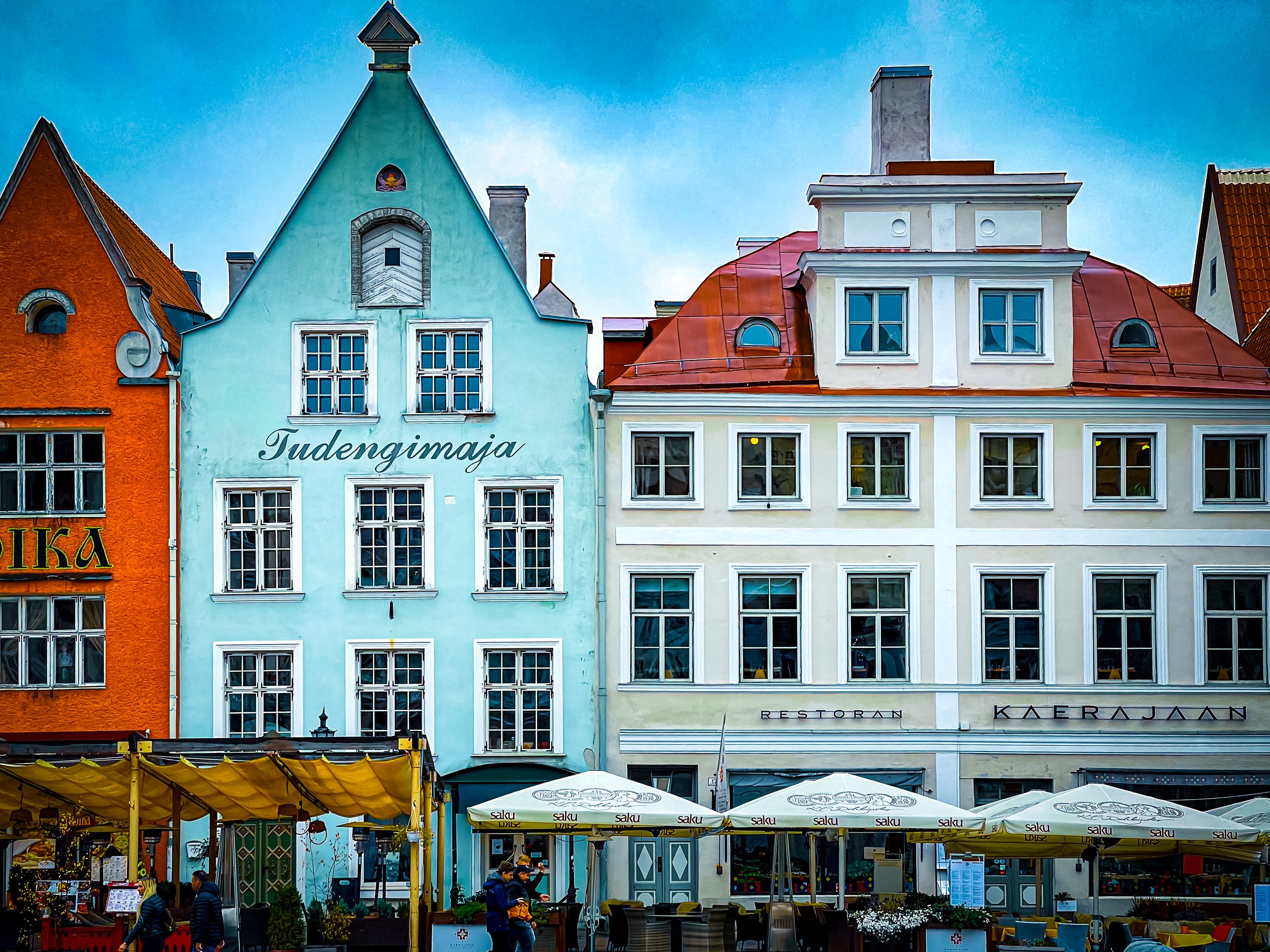 Colorful row of buildings with the words 'Tudengmaja' on a light blue building, outdoor seating with umbrellas, and people walking in a lively town setting.