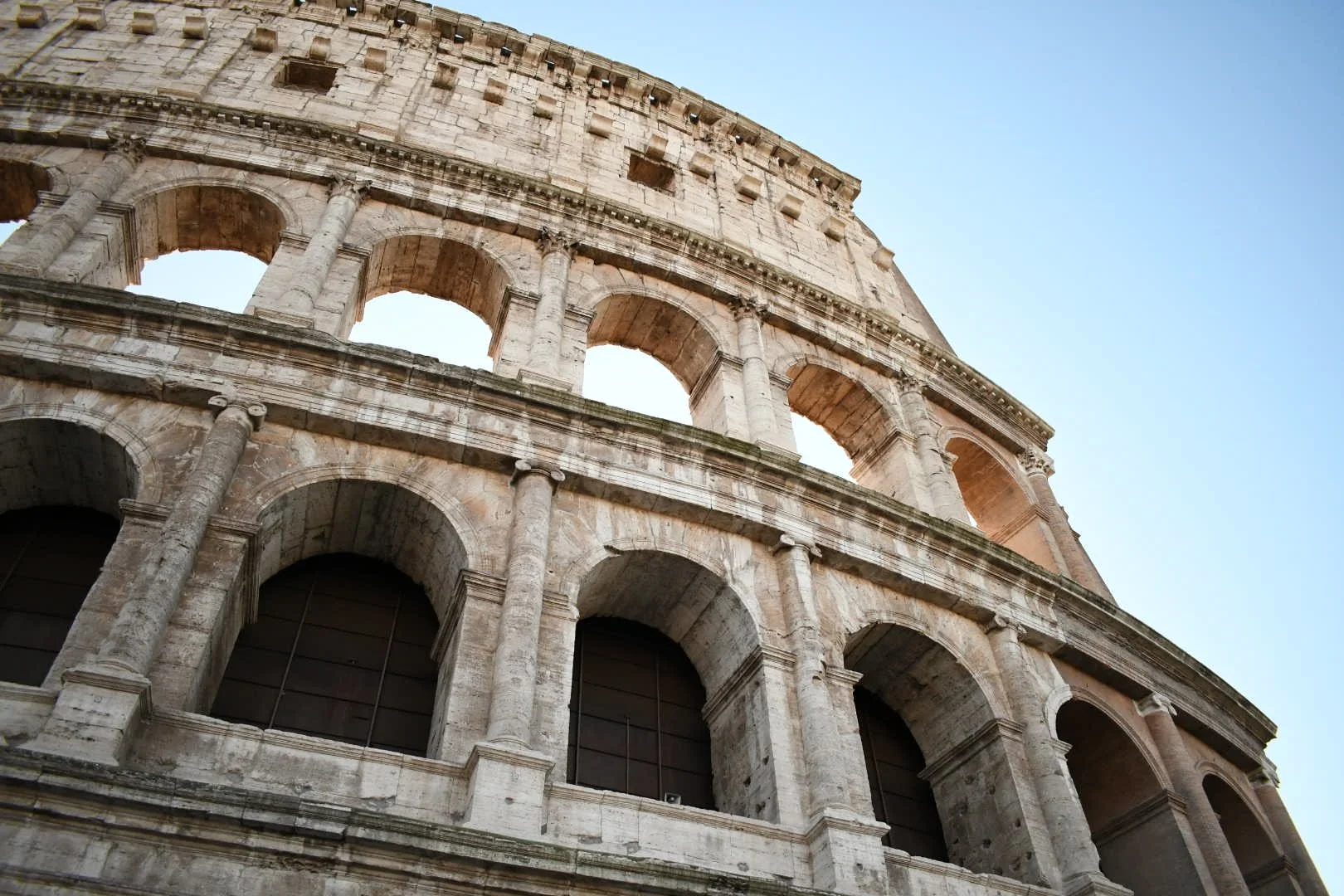 Close-up of the exterior of the ancient Roman Colosseum showing multiple levels of arches and stonework against a blue sky.