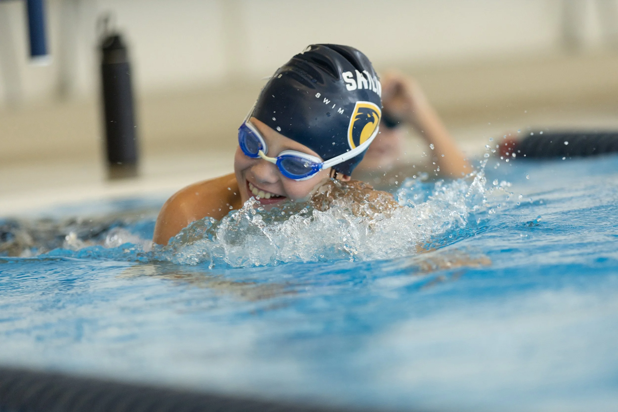 Child wearing a swim cap and goggles swimming in a pool with water splashing around.