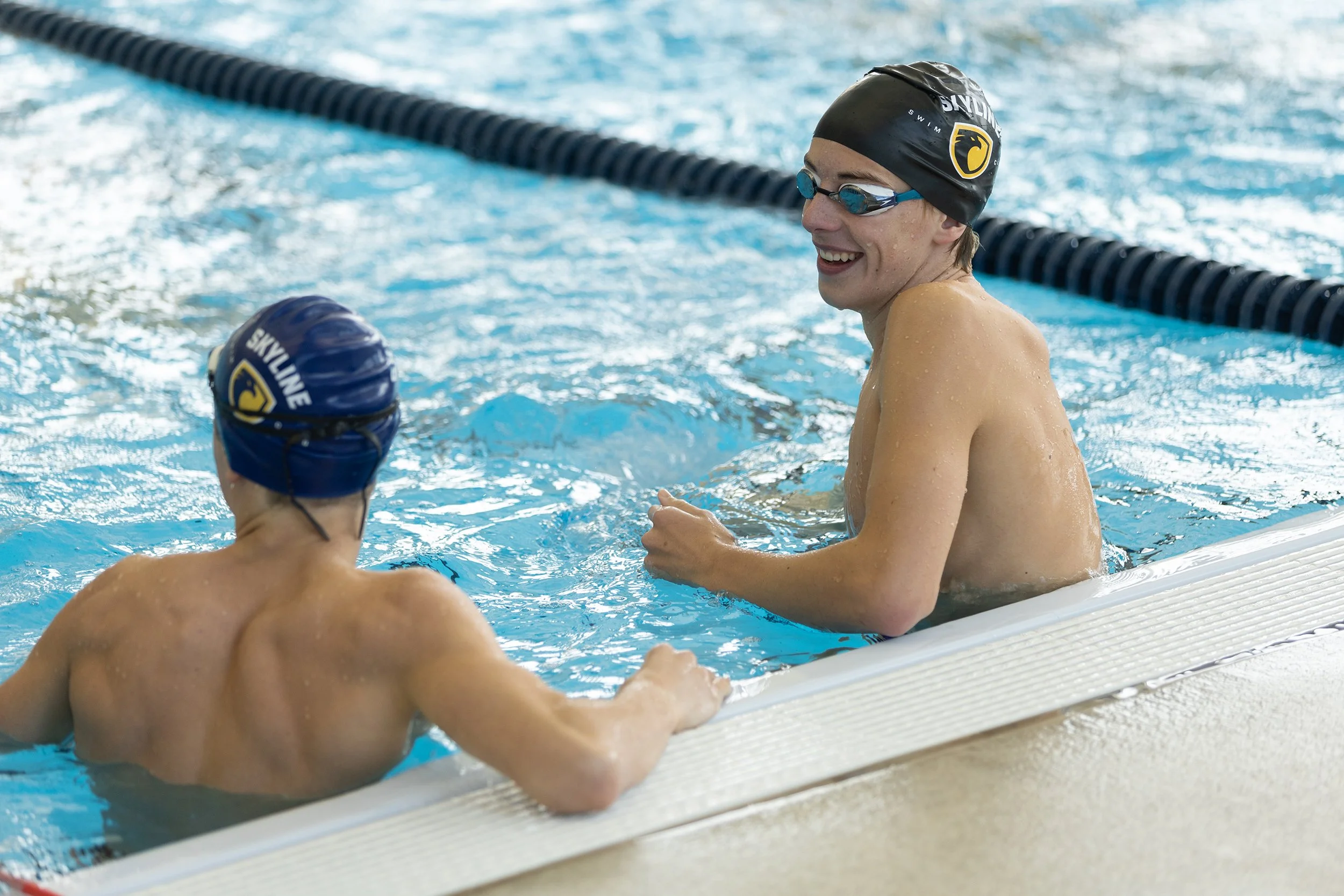 Smiling swimmer talking to a child swimmer at the edge of a swimming pool, both wearing swim caps and goggles.