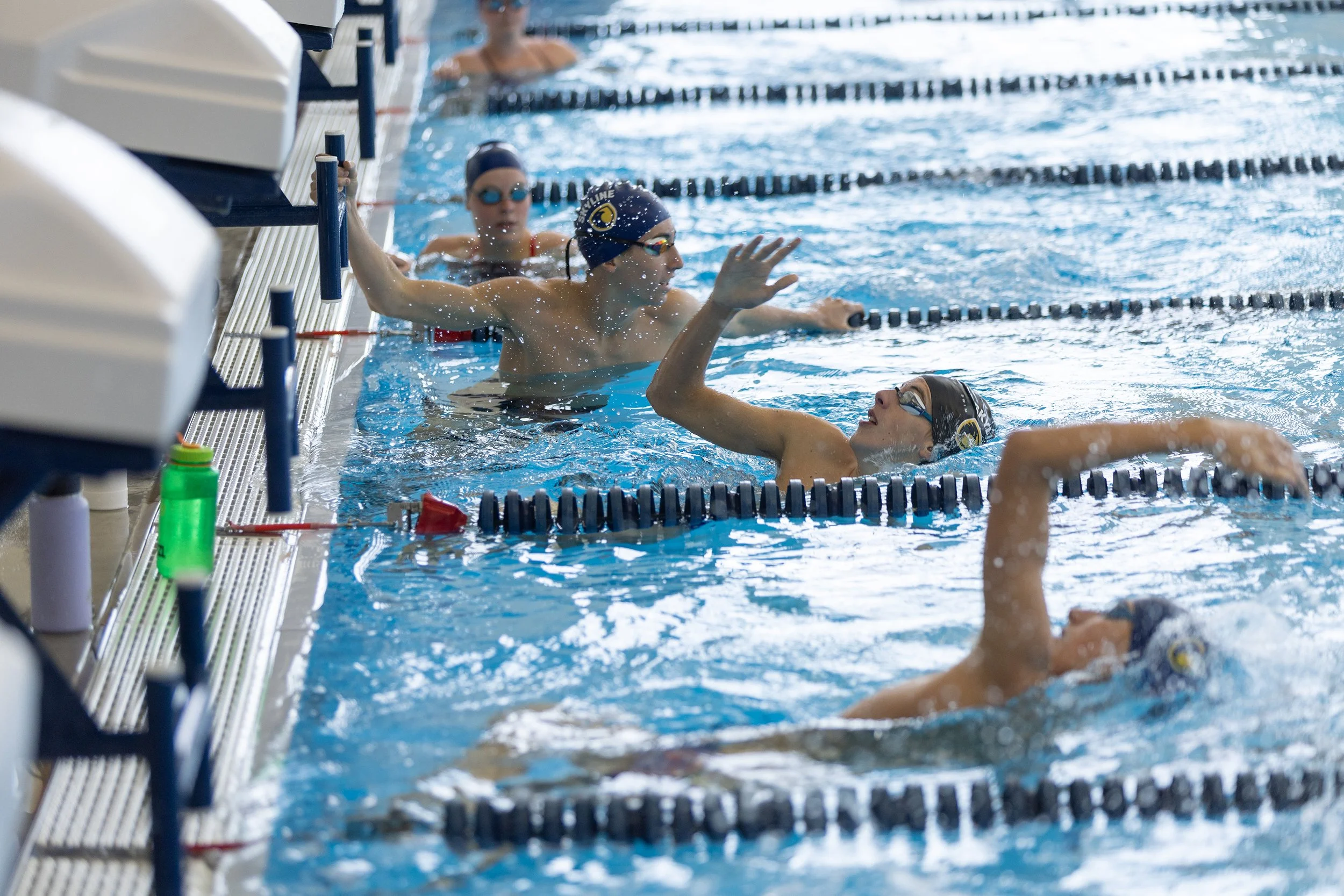 Swimmers at the edge of a swimming pool during swim practice, wearing swim caps and goggles, with lane dividers visible in the water.