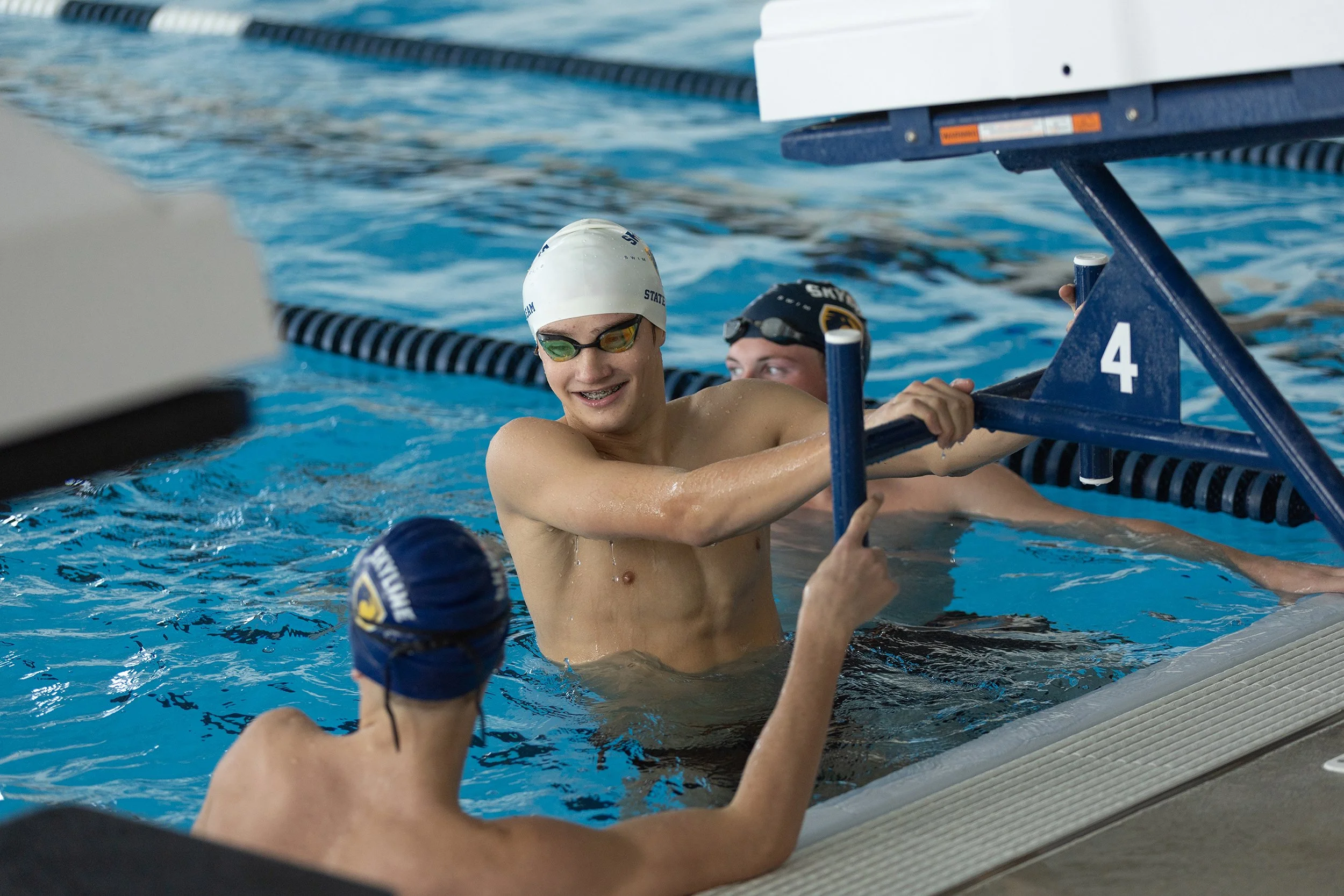 Swimmers in a pool, wearing swim caps and goggles, a young man smiling and holding onto the starting block.