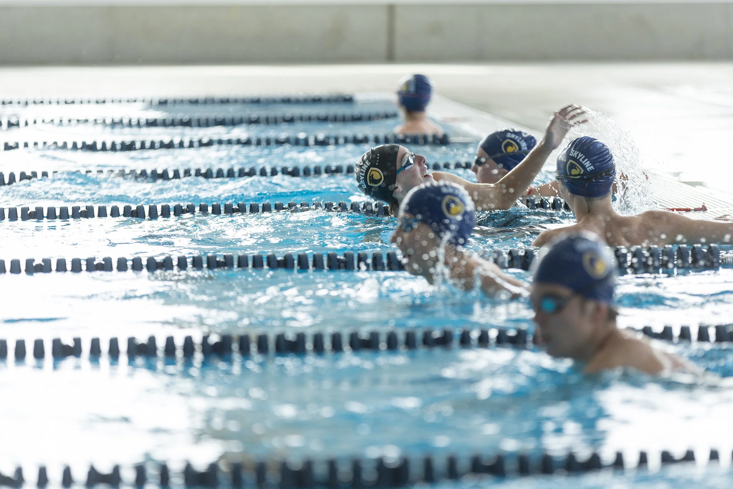 Swimmers in a swimming pool near the wall, wearing blue swim caps and goggles, engaged in a swimming practice.