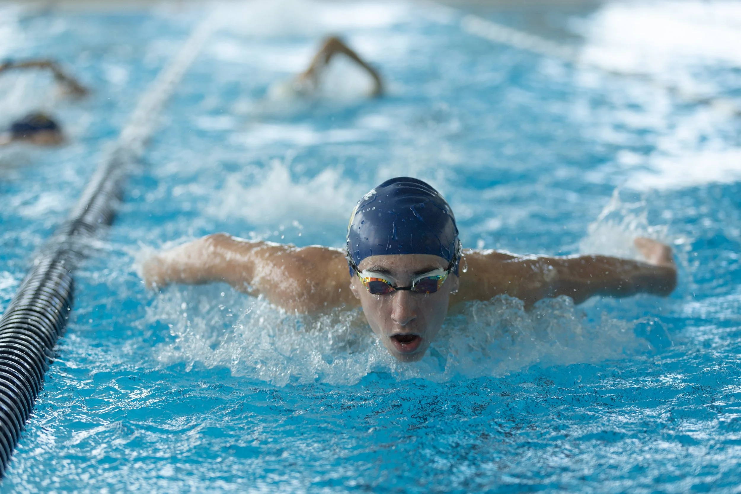 Swimmer wearing a blue swim cap and goggles swimming in a pool, viewed from the front, with water splashing around.