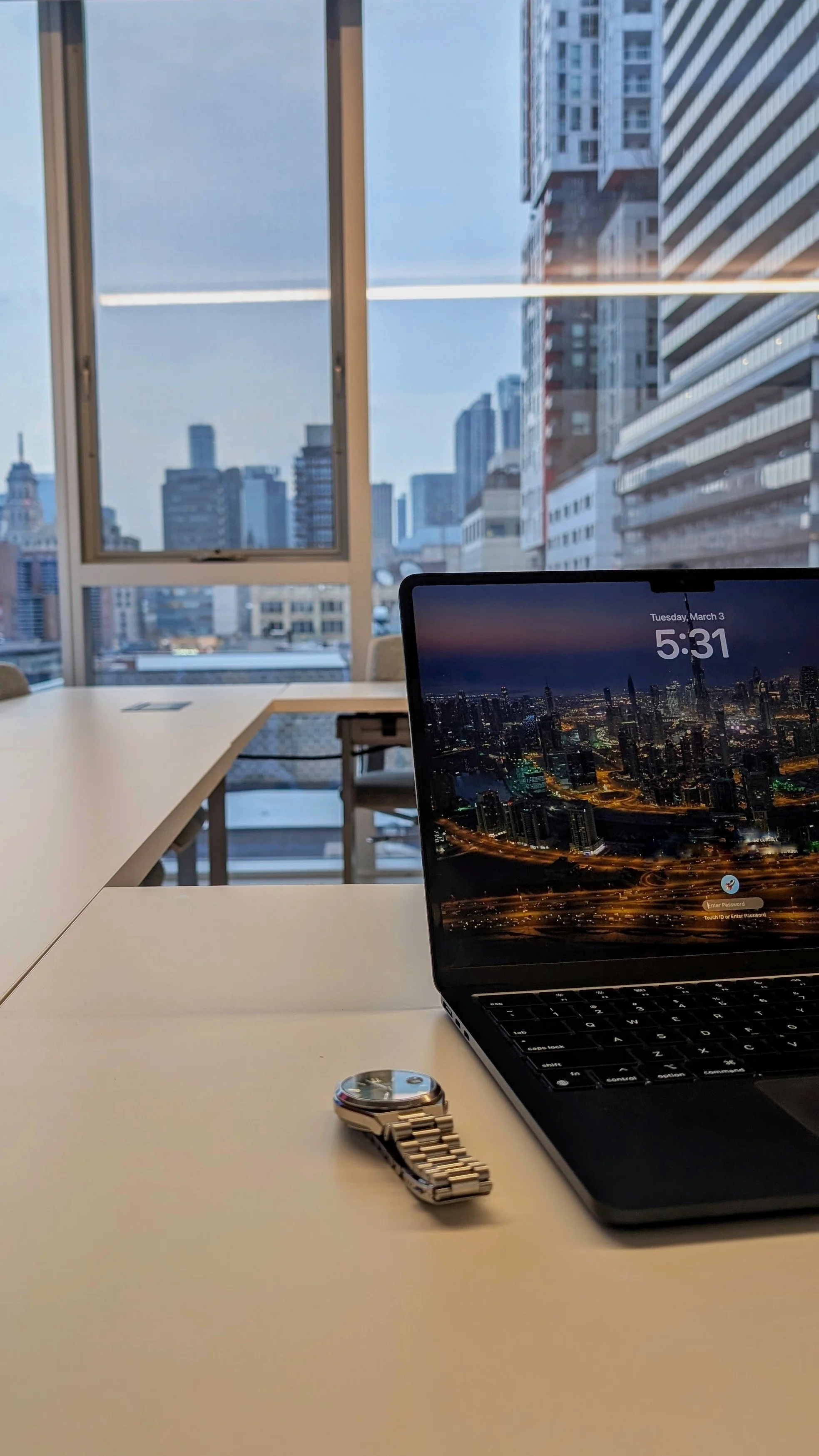 Office workspace with a laptop displaying a city skyline at night, a silver wristwatch, and a round object on a white table, with city buildings visible through large windows in the background.