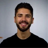 A young man with dark hair, a beard, and a mustache, smiling, wearing a black shirt, against a plain light gray background.