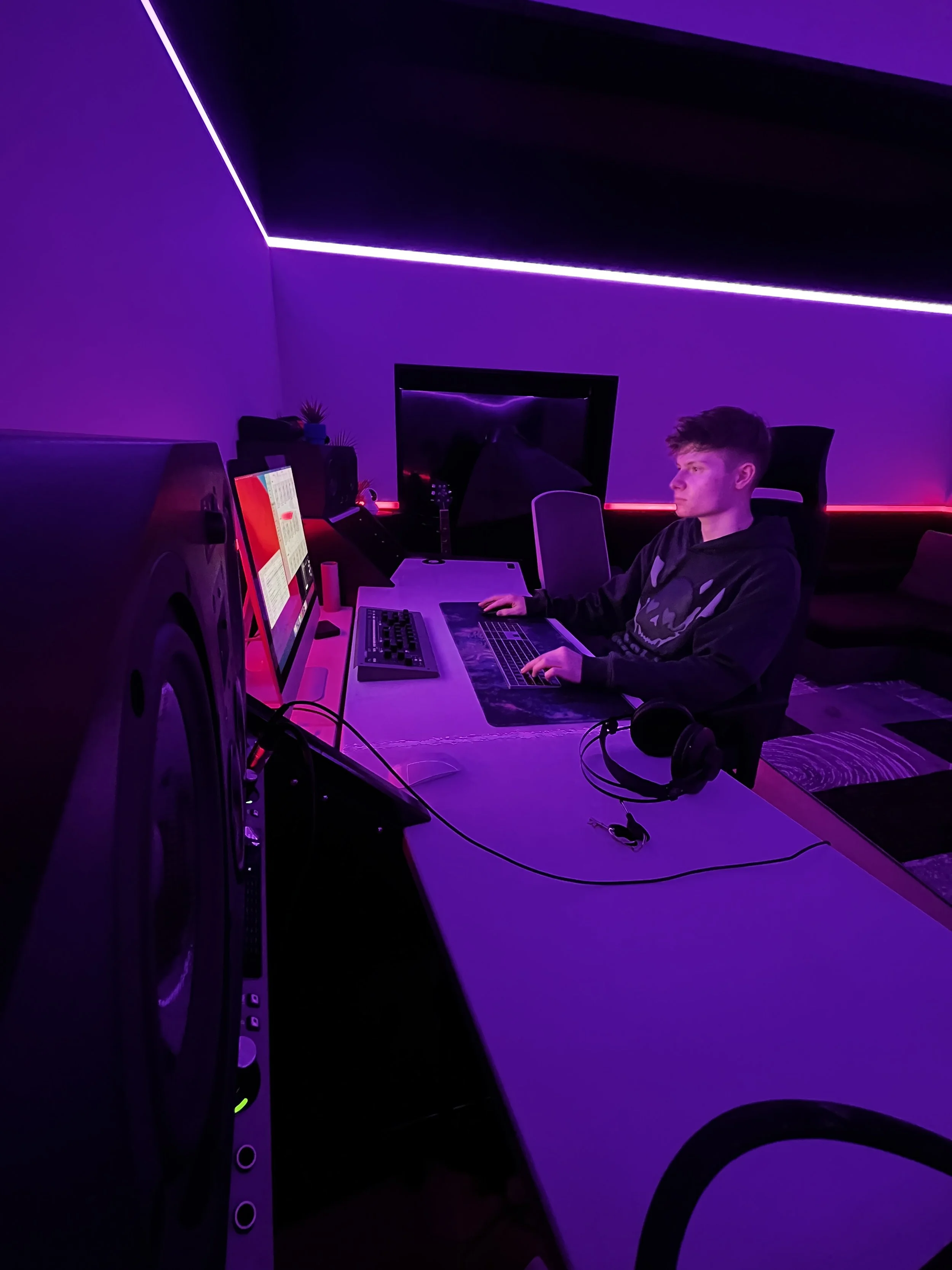 Young man working at a desk with dual monitors illuminated by purple and pink neon lighting in a modern, dimly lit room.