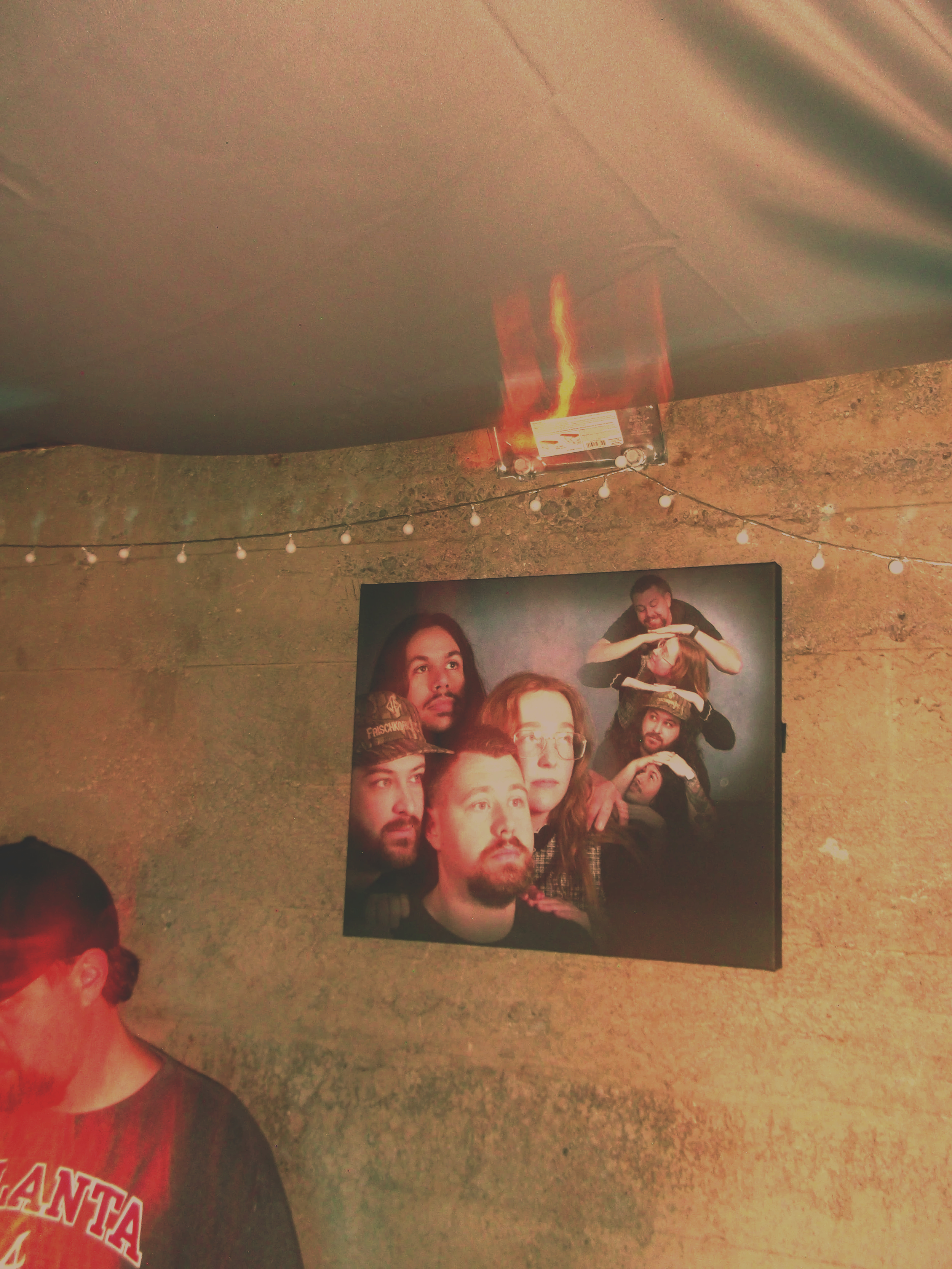 The image shows a group photo of several people with diverse facial expressions, positioned against a dark background with some lighting effects. The top part of the image features a ceiling with a visible tile pattern and a hanging light fixture, while the lower part includes a poster or photograph on the wall and some people in the foreground.