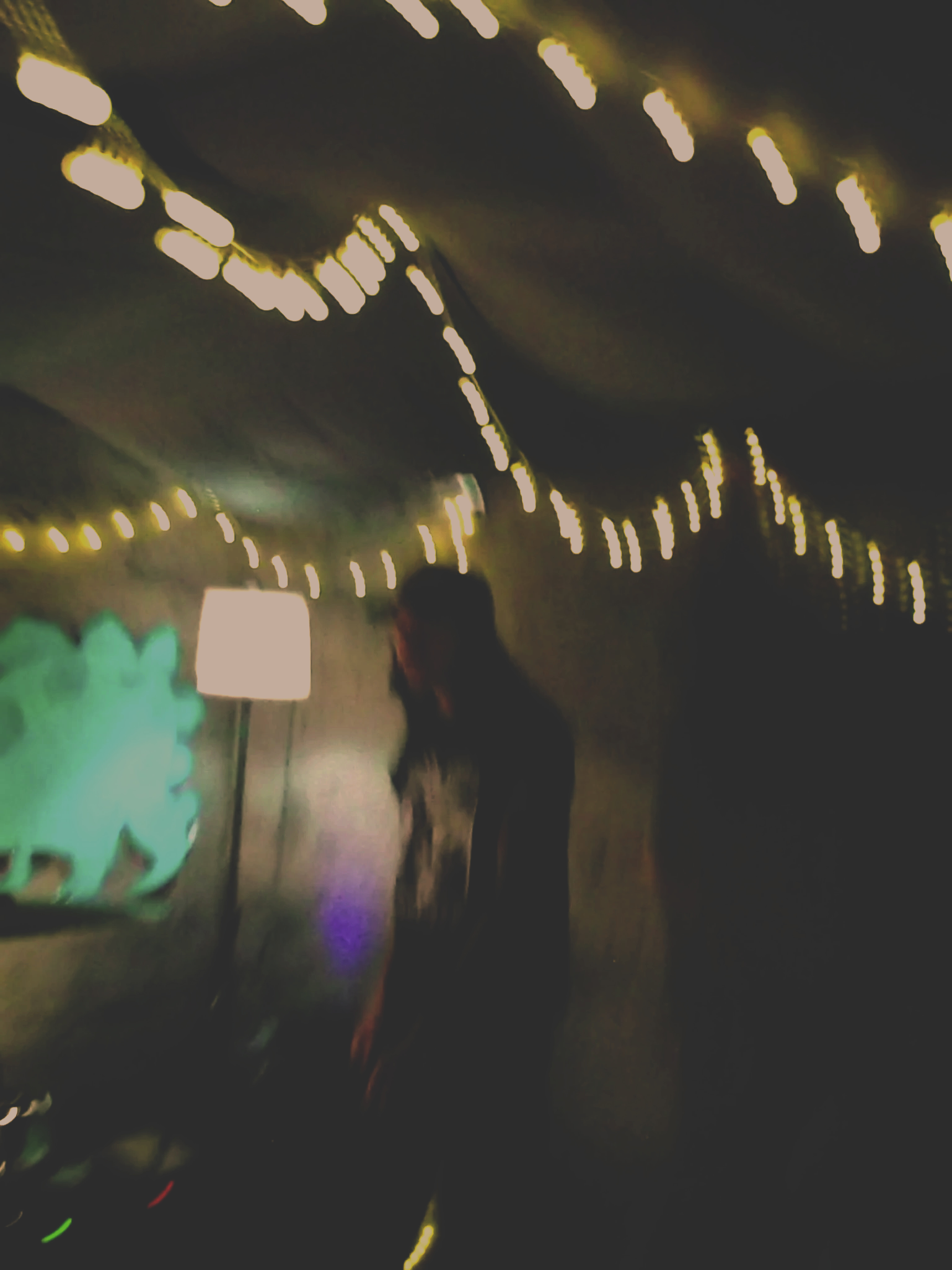 Blurred image of a person standing in a dark room decorated with string lights, appears to be festive or party setting.
