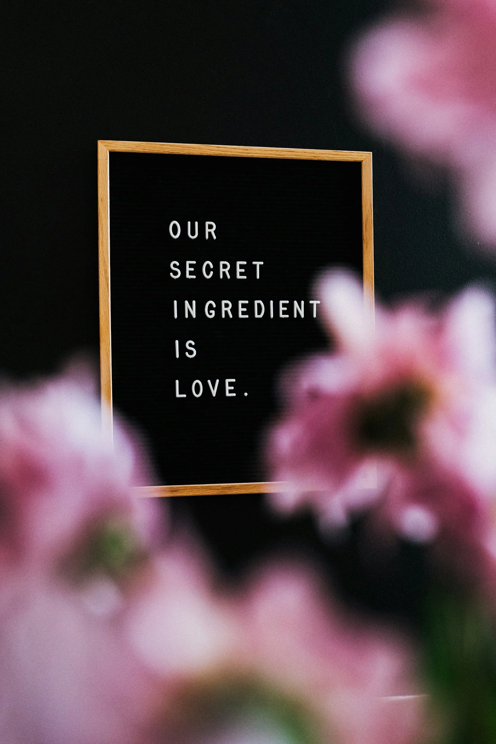 A black letter board with wooden frame displaying the words "OUR SECRET INGREDIENT IS LOVE" in white letters, blurred pink flowers in the foreground, and a dark background.