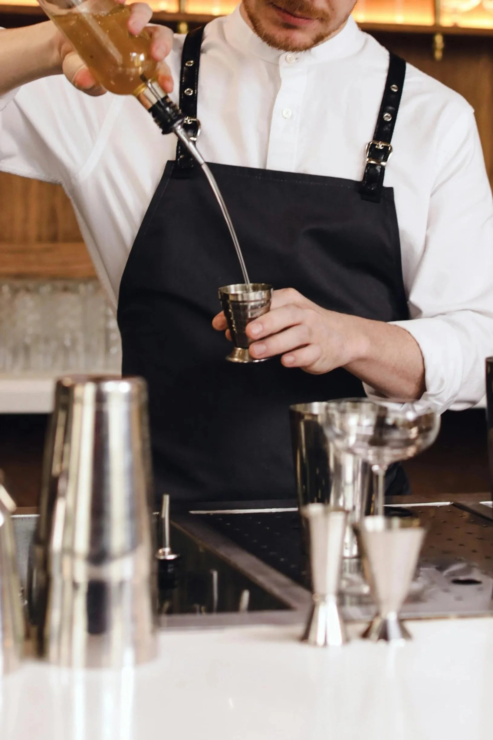 Bartender pouring a drink into a jigger at a bar, with bar tools in the foreground.