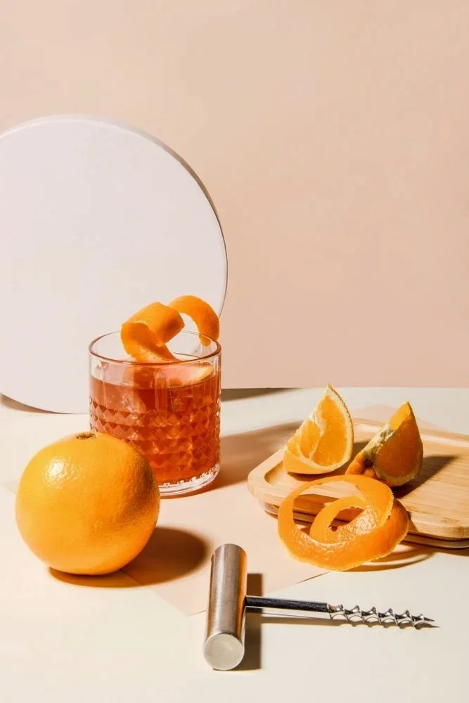 An old fashioned cocktail with orange peel garnish, a whole grapefruit, orange slices, orange peels on a wooden tray, and a cork screw opener tool on a light-colored surface with a plain background.
