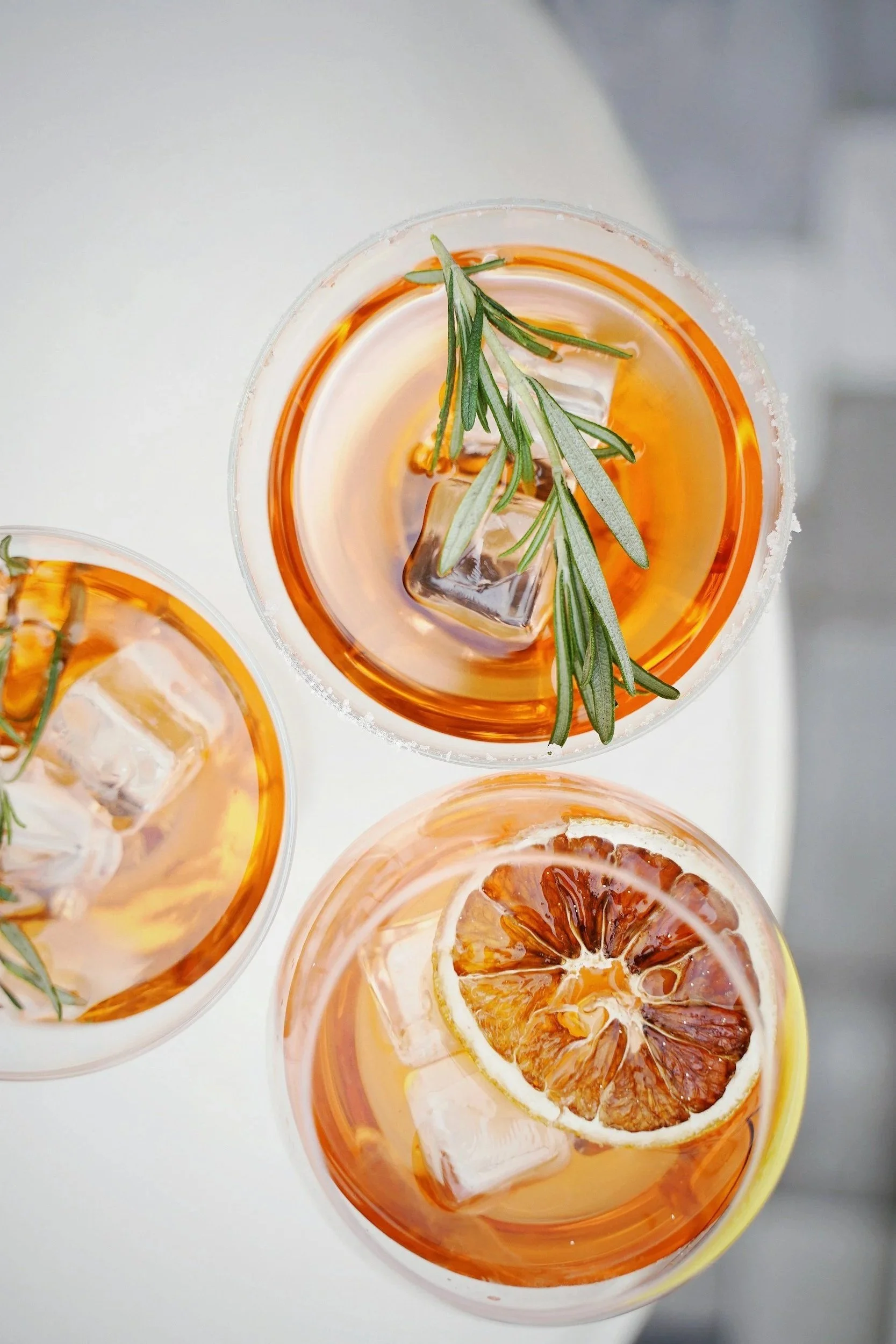 Close-up of three glassses of orange-colored cocktails with a sprig of fresh rosemary and a large ice cube, garnished with a slice of dried blood orange.