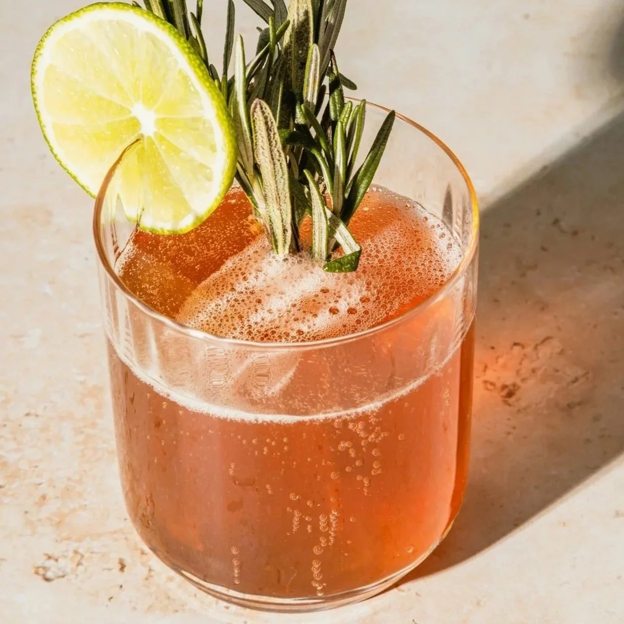 A beautiful cocktail garnished with a slice of lemon and sprigs of rosemary on a light-colored surface.