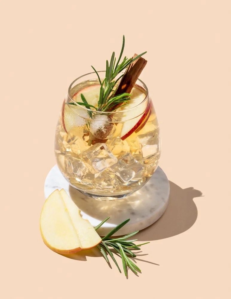 An artisanal apple cider cocktail with ice, garnished with apple slices, a cinnamon stick, and a sprig of rosemary, placed on a white marble coaster with an apple slice and rosemary sprig beside it.