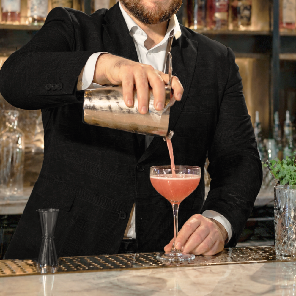 A bartender in a black suit and white shirt is pouring a pink lady gin based cocktail into a coupe cocktail glass. In the background there is a bar with liquor bottles on shelves.