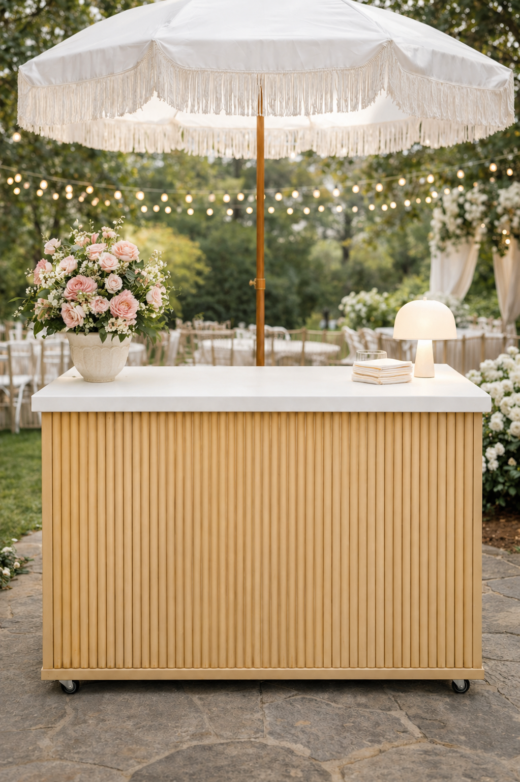 Outdoor event bar setup with a wooden bar, large pink and white flower arrangement, white mini table lamp, and white umbrella, surrounded by string lights and decorated tables.